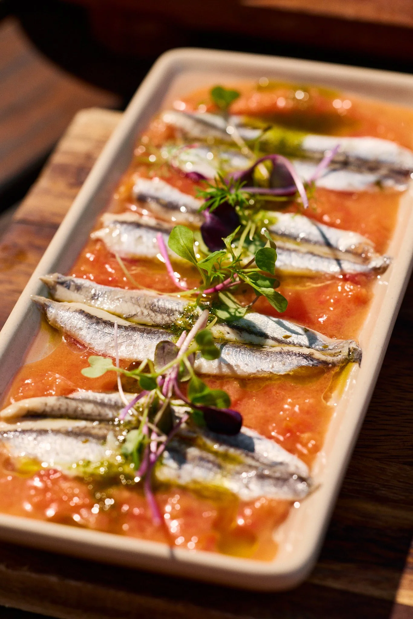 Sardines garnished with microgreens and drizzled with oil, served on a bed of tomato sauce in a white rectangular dish.