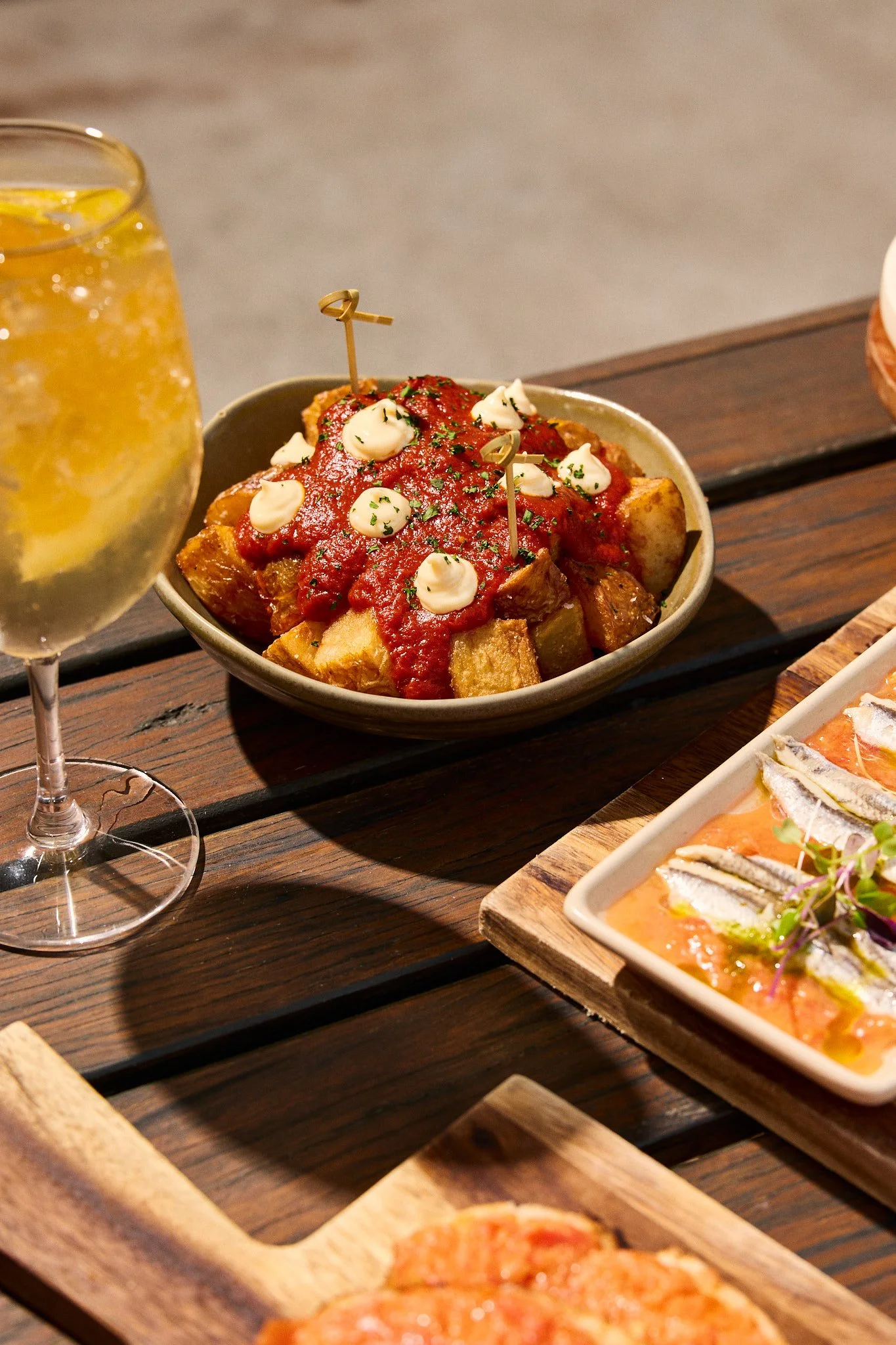 A bowl of potato wedges topped with tomato sauce, mayonnaise, and herbs on a wooden table, with a glass of sparkling lemon water nearby and other dishes.