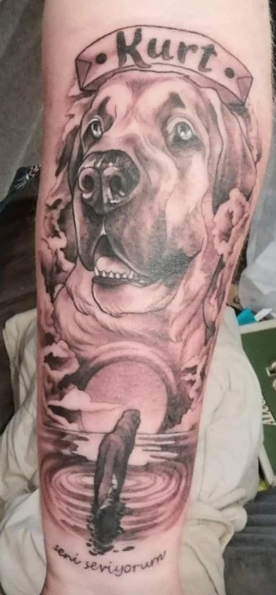 Tattoo of a large dog’s face with a banner labeled 'Kurt' above it, a person walking into a sunset over water at the bottom, and words in a foreign language below.