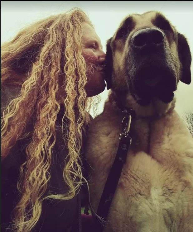 Woman with long, curly blonde hair giving a kiss on the nose to a large Mastiff dog with a light-colored coat and black markings on its face outdoors.