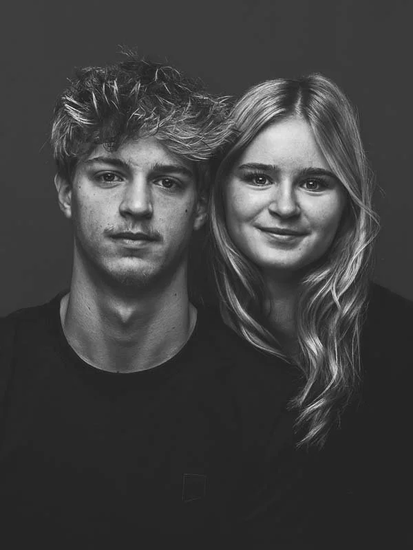 Zwart-wit foto van een jonge man en een jonge vrouw, zittend dicht bij elkaar, glimlachend, met een donkere achtergrond.
