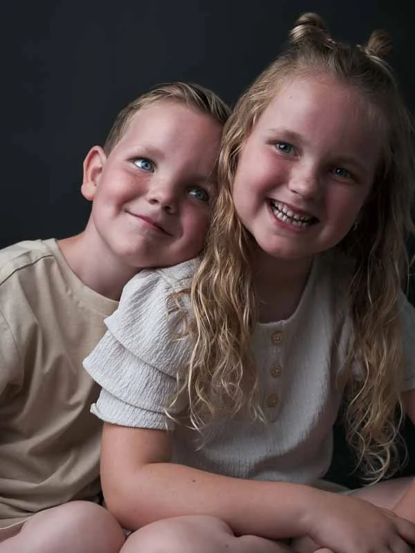 Twee jonge kinderen, een jongen en een meisje, glimlachen en zitten dicht bij elkaar tegen een donkere achtergrond.
