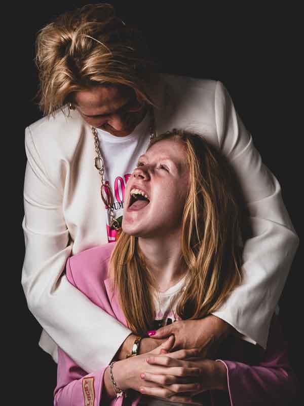 Twee vrouwen, een oudere en een jongere, lachen en omhelzen elkaar liefdevol op een donkere achtergrond.