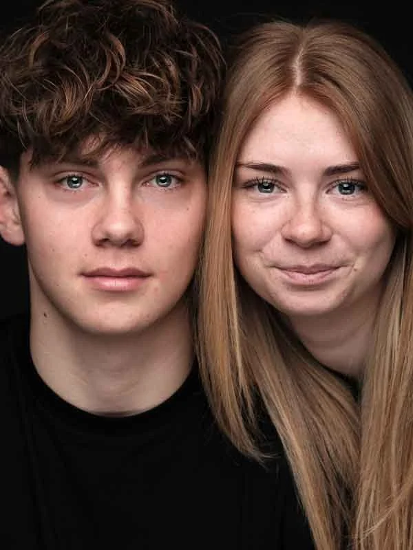 Twee jonge mensen, een man met krullig haar en een vrouw met recht haar, kijken naar de camera. Ze zitten dicht bij elkaar tegen een zwarte achtergrond.