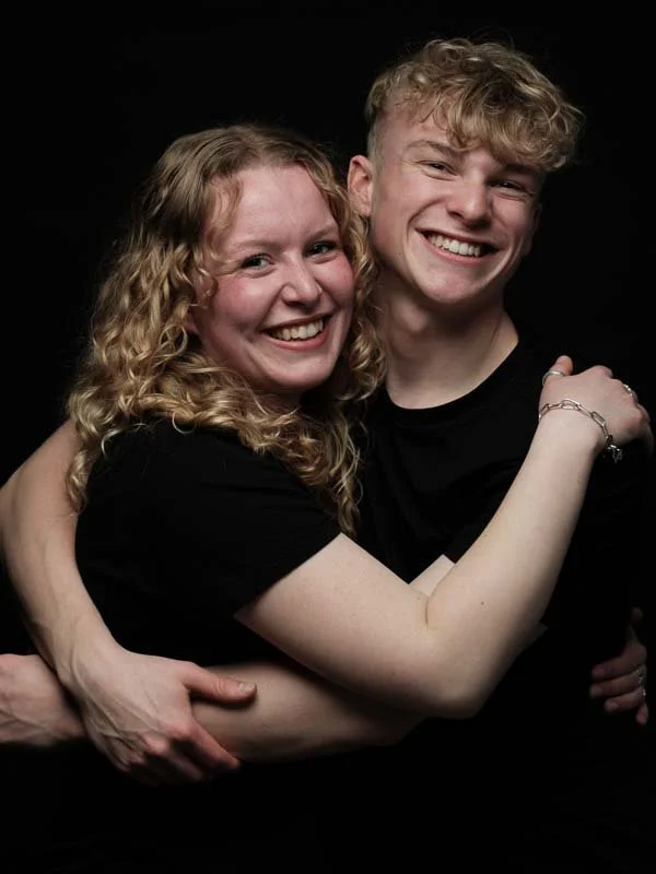 Twee jonge mensen, een man en een vrouw, die lachen en elkaar omarmen tegen een zwarte achtergrond.