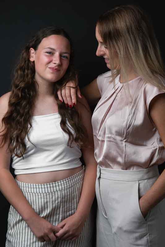 Twee vrouwen staan samen, een glimlacht en kijkt naar de camera, de andere kijkt naar haar en glimlacht, beide dragen lichte kleding, tegen een donkere achtergrond.