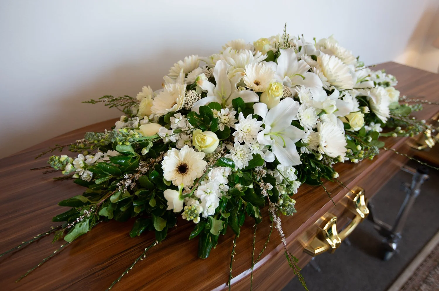 White floral arrangement on a polished wooden casket with brass handles, including lilies, roses, gerbera daisies, and greenery.