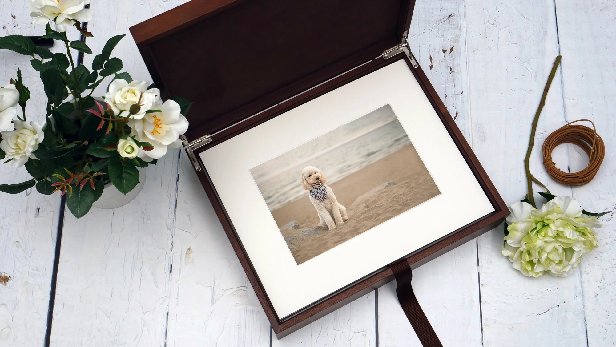A framed photo of a white dog sitting on the beach, surrounded by white and green flowers, with some craft materials on a white wooden surface.