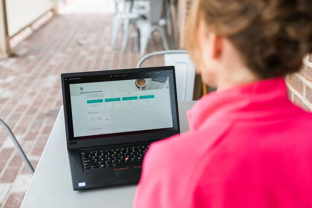 a woman in a pink jacket sitting at an outdoor coffee shop and sharing her computer screen with a client