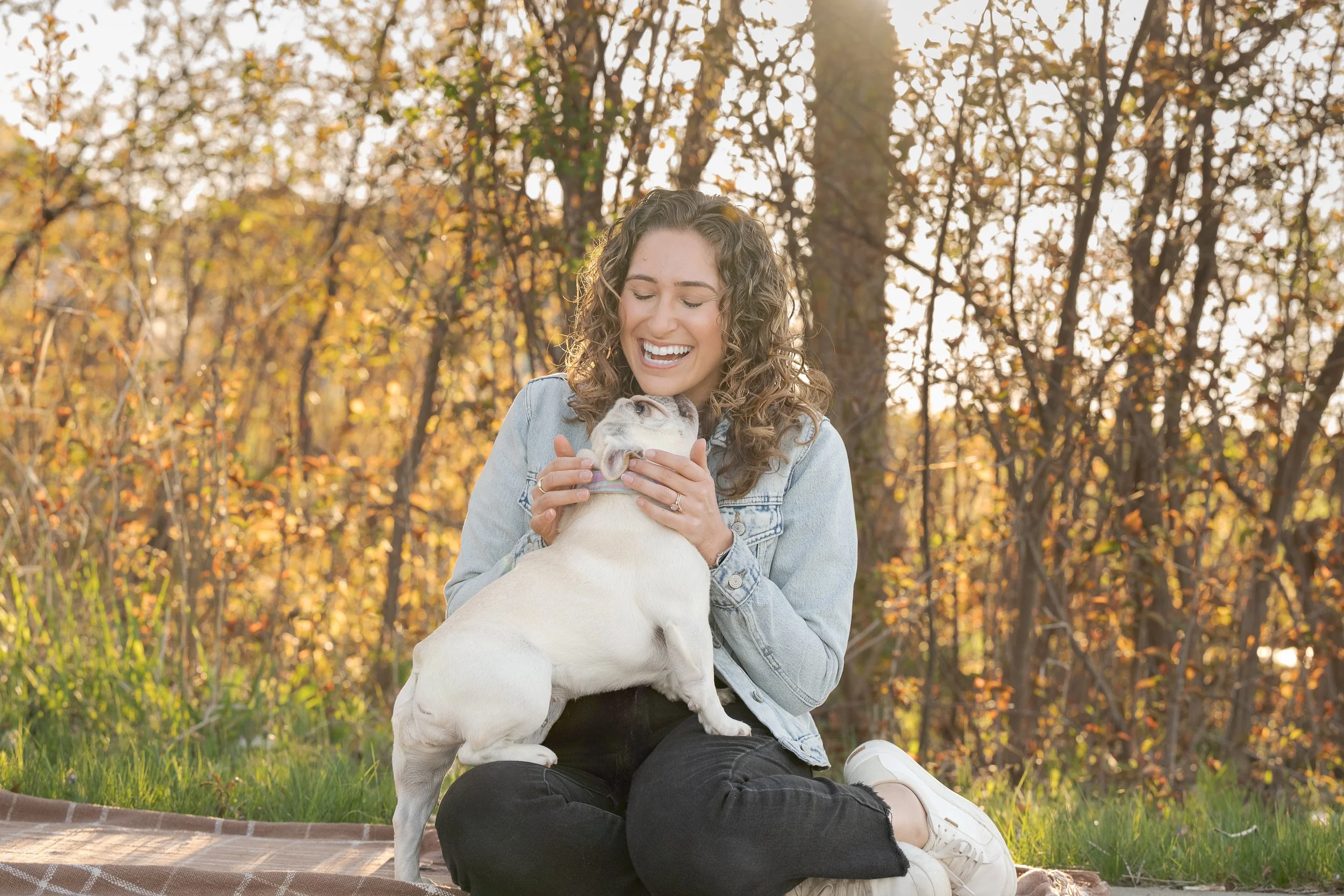 A woman with curly hair wearing a denim jacket and black pants is sitting outdoors on a blanket in a park during autumn, holding a small dog and smiling happily.