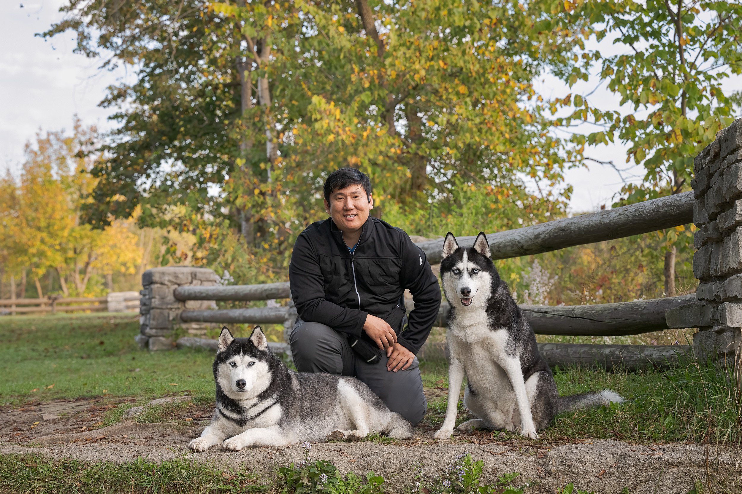 A man with two Siberian Huskies, one lying down and one sitting, outdoors in a park during autumn with trees in the background.