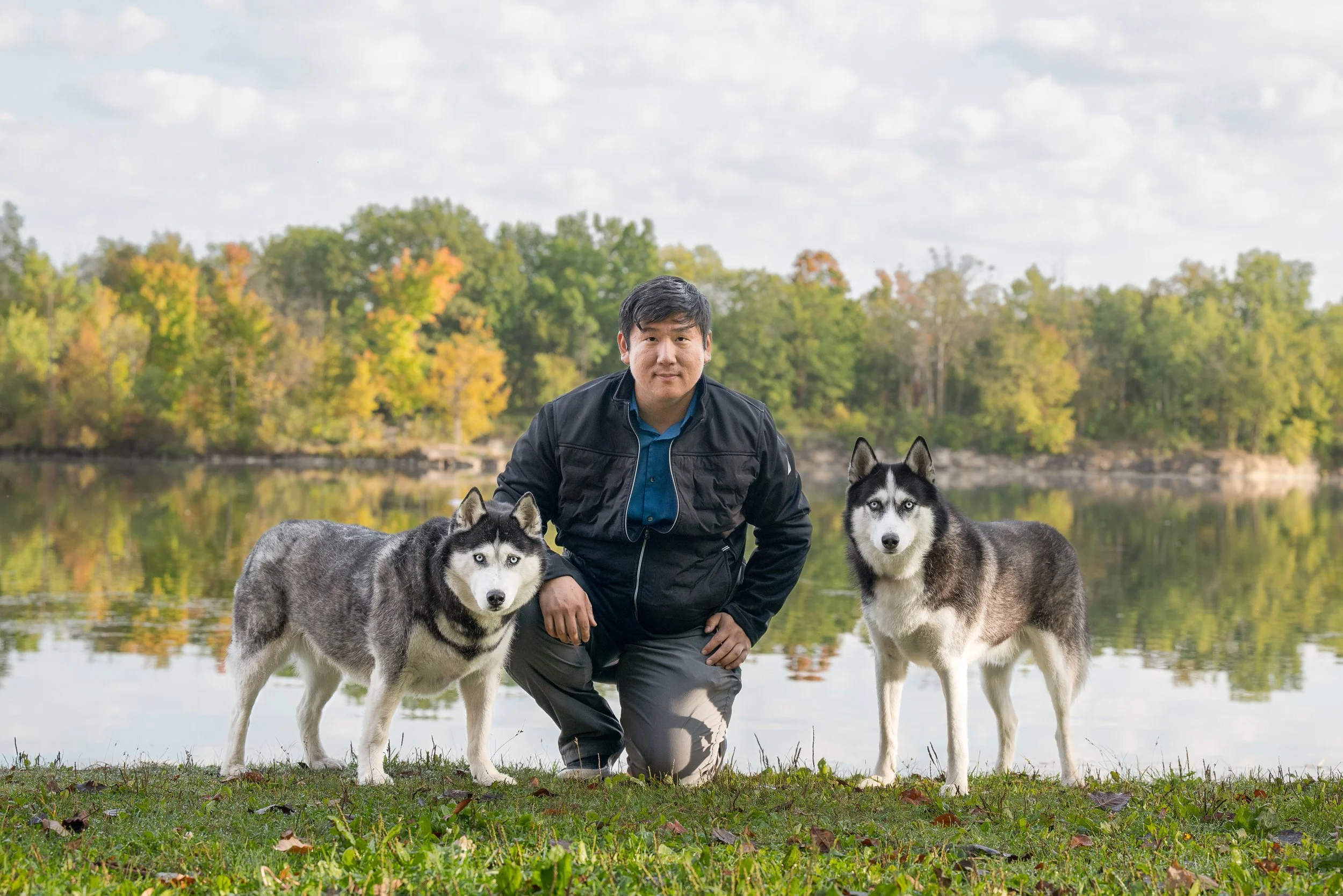 A man kneeling on grass with two huskies near a lake, surrounded by trees with autumn foliage.
