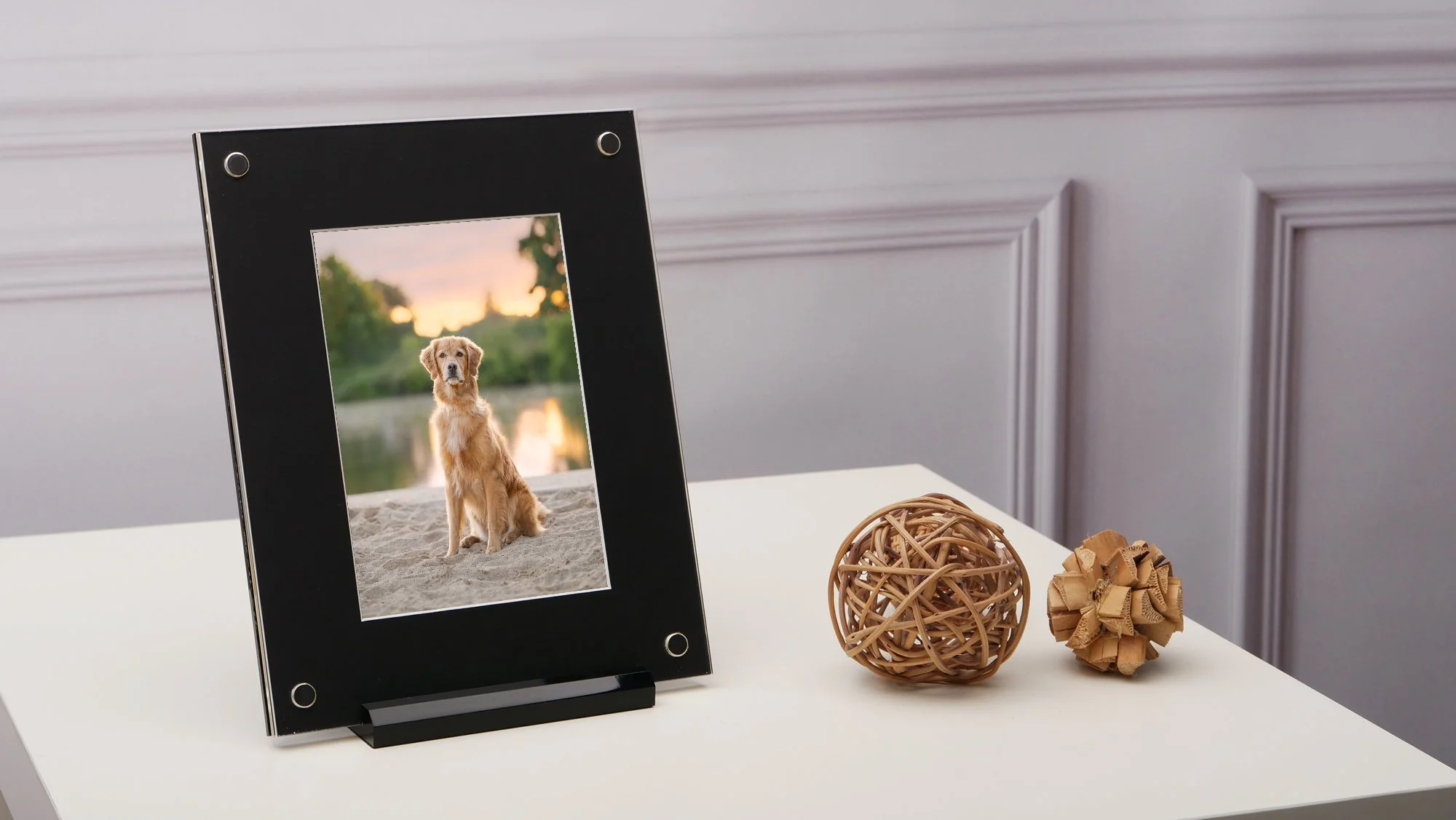 A black picture frame with a photo of a golden retriever dog sitting on sand near a river at sunset. Two decorative woven balls made of rattan are on a white surface next to the frame.