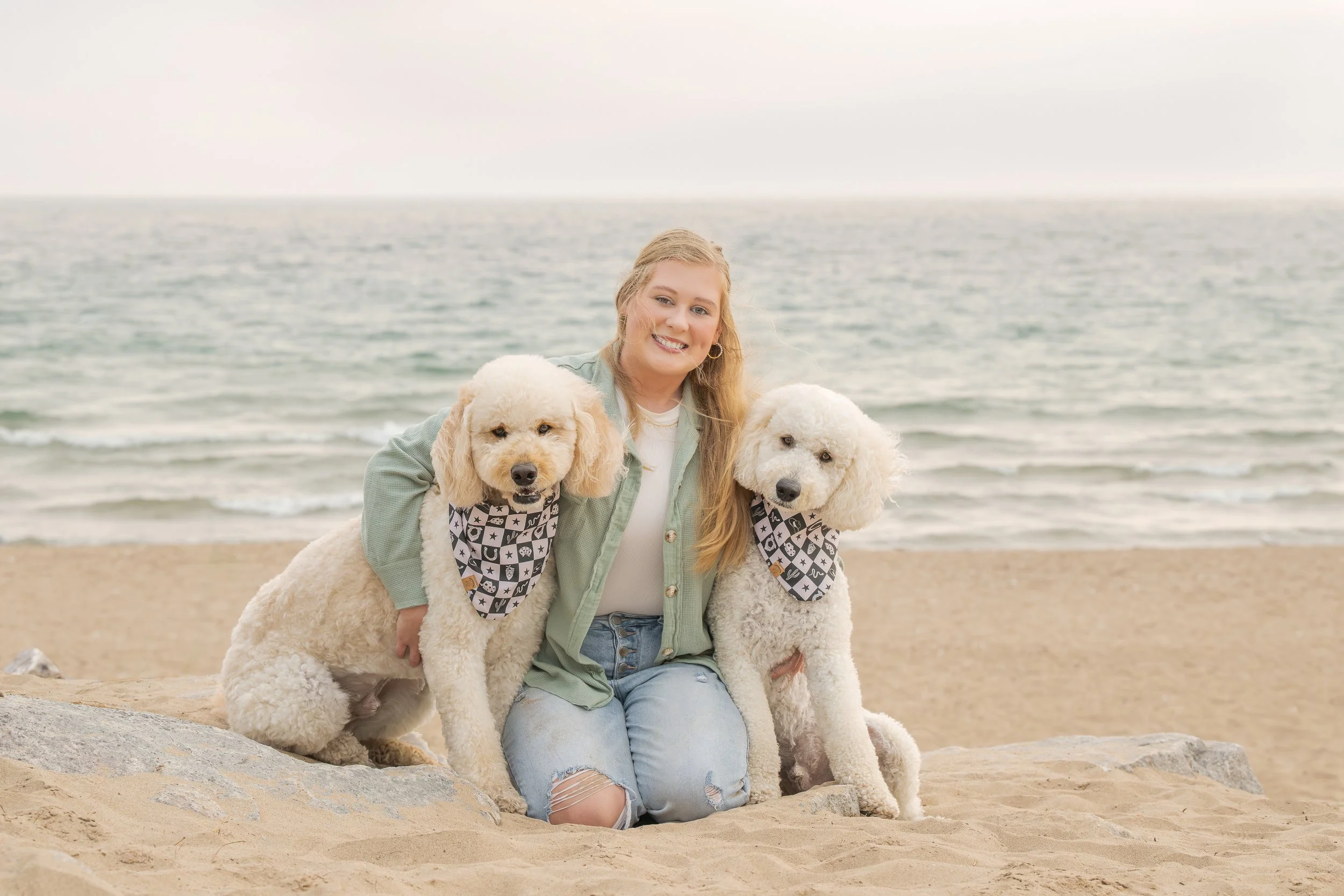 A lovely young lady with long blonde hair sitting on the beach at Lake Michigan in Oak Creek, Wisconsin with her two cute Golden Doodle Dogs.