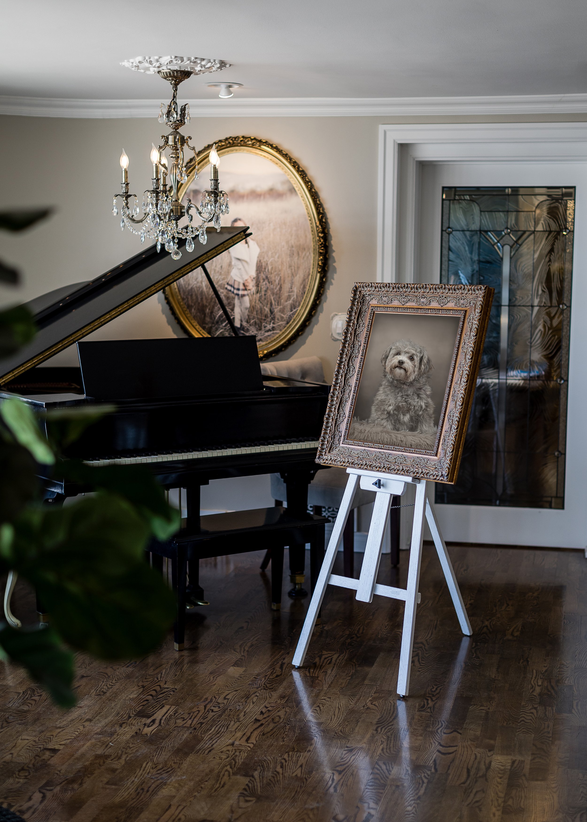 An interior living room with a grand piano, a chandelier, a framed portrait of a dog on an easel, and a large framed painting on the wall.