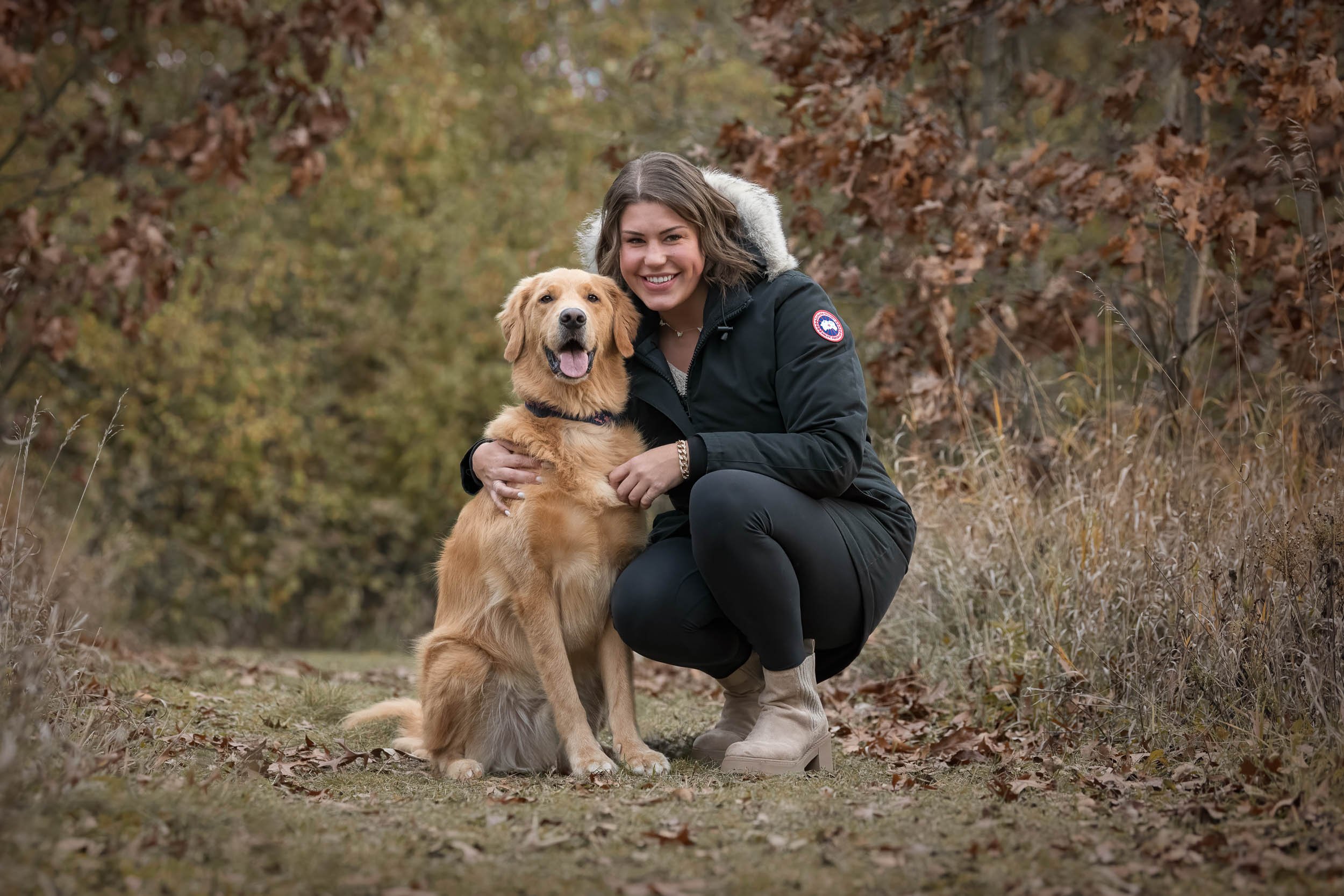 A woman crouching outdoors with a golden retriever dog in a wooded area during autumn.