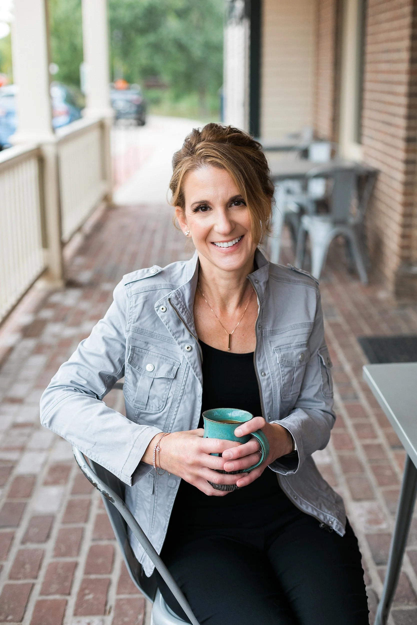 a woman in a grey jacket and black pants sitting at an outdoor pet-friendly coffee shop holding a coffee mug
