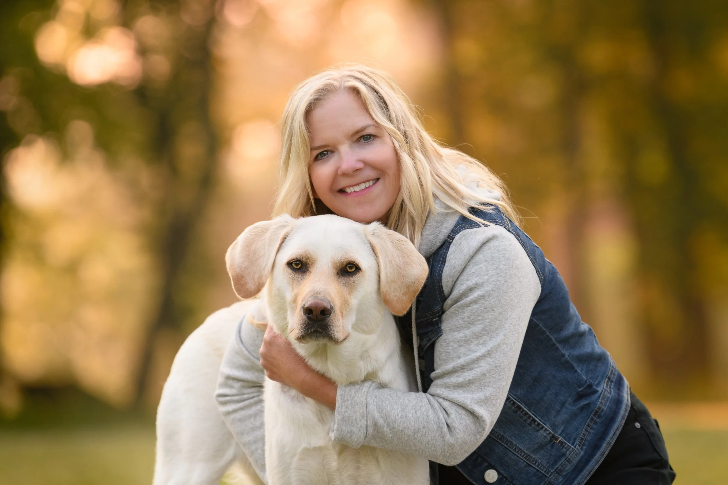 A woman smiling and holding a light-colored Labrador Retriever puppy outdoors in a park during autumn, with golden trees in the background.