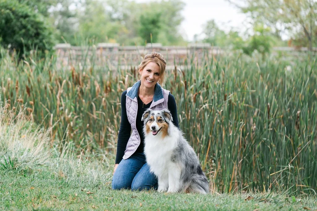 Photo of a woman in a puple vest with her australian shepherd in front of a bridge