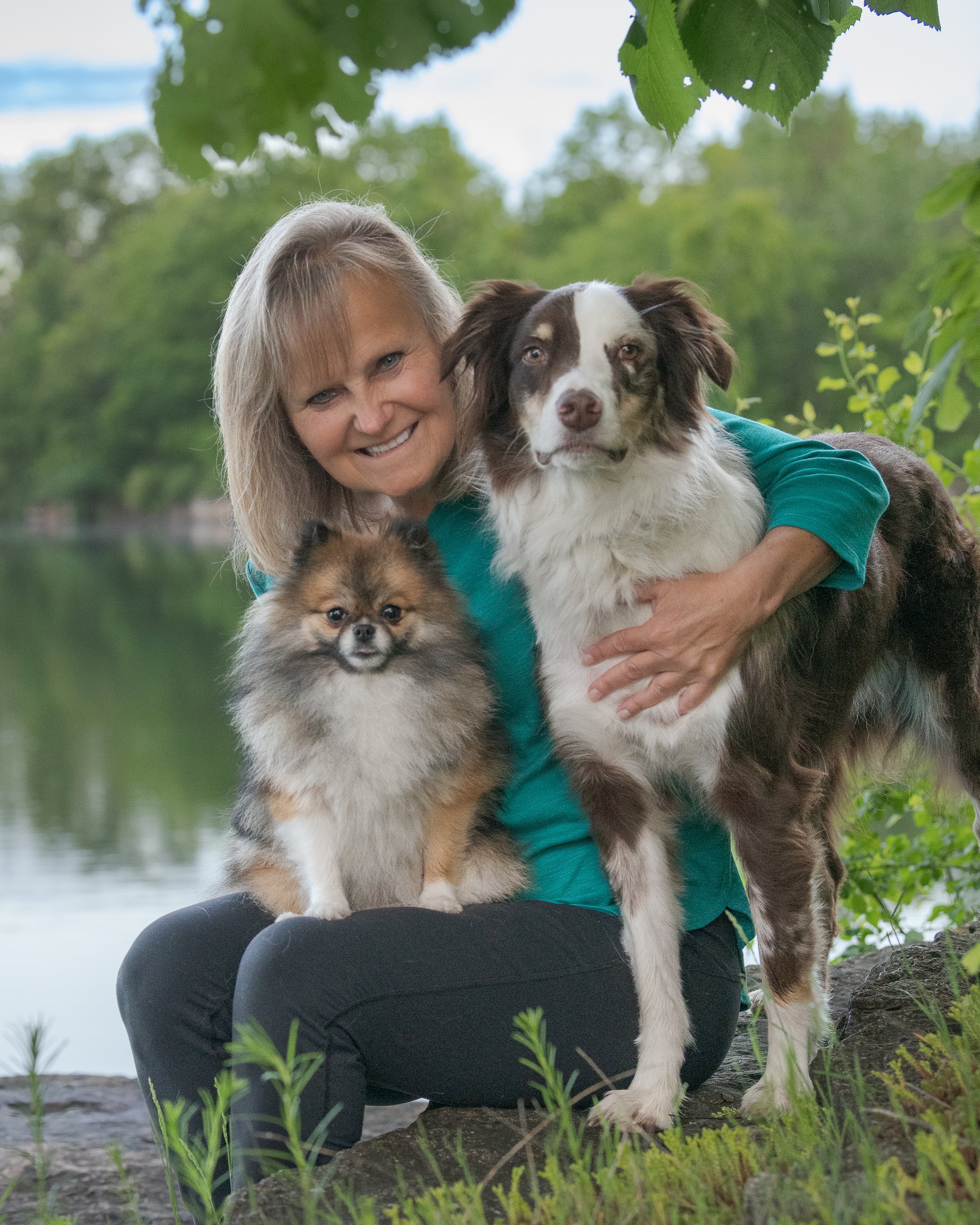 A woman with two dogs sitting outdoors near a body of water, with green trees in the background.