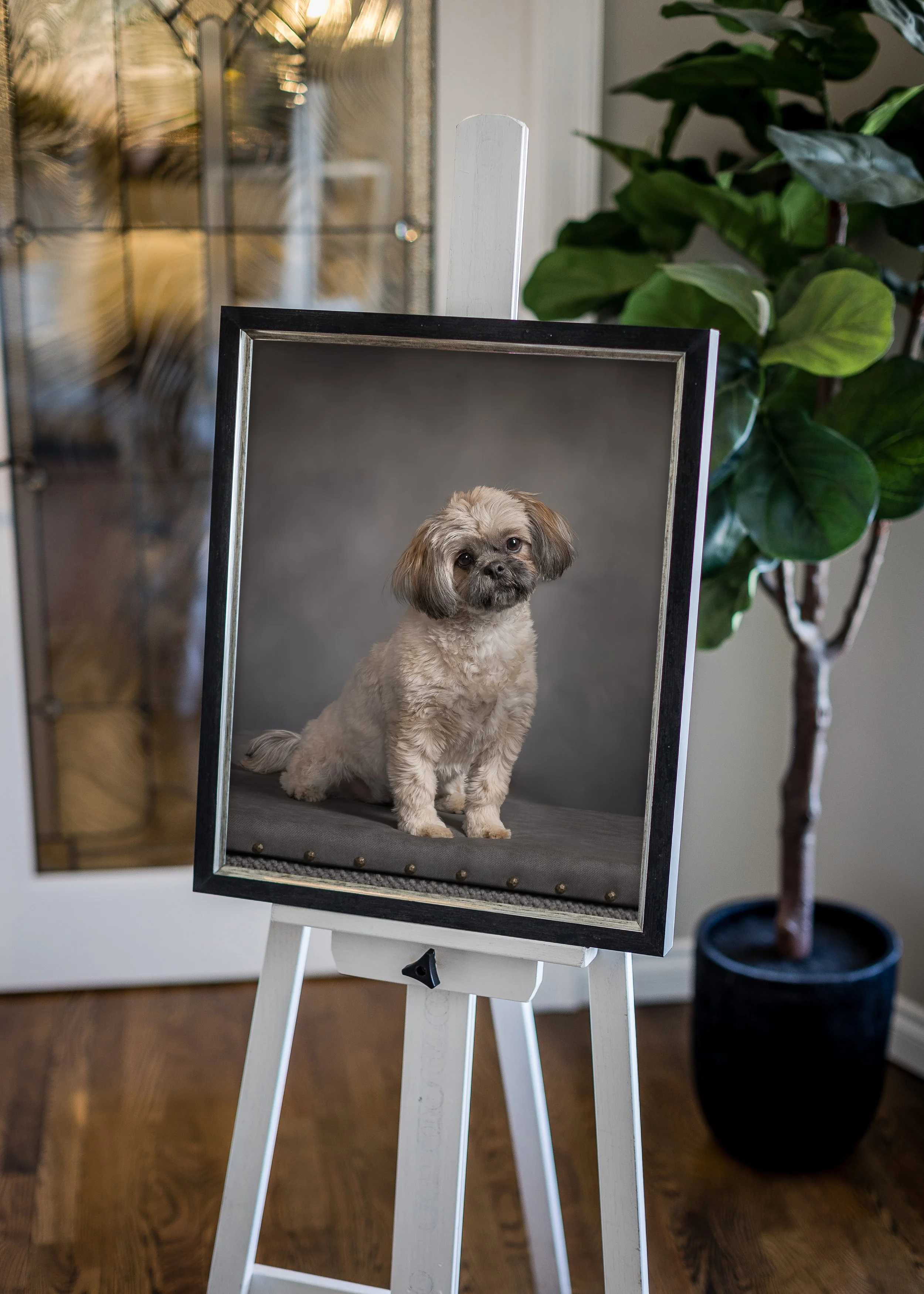 A dog portrait on an easel, featuring a small, fluffy dog with floppy ears and sad eyes, against a gray background, indoors with a plant and a door in the background.