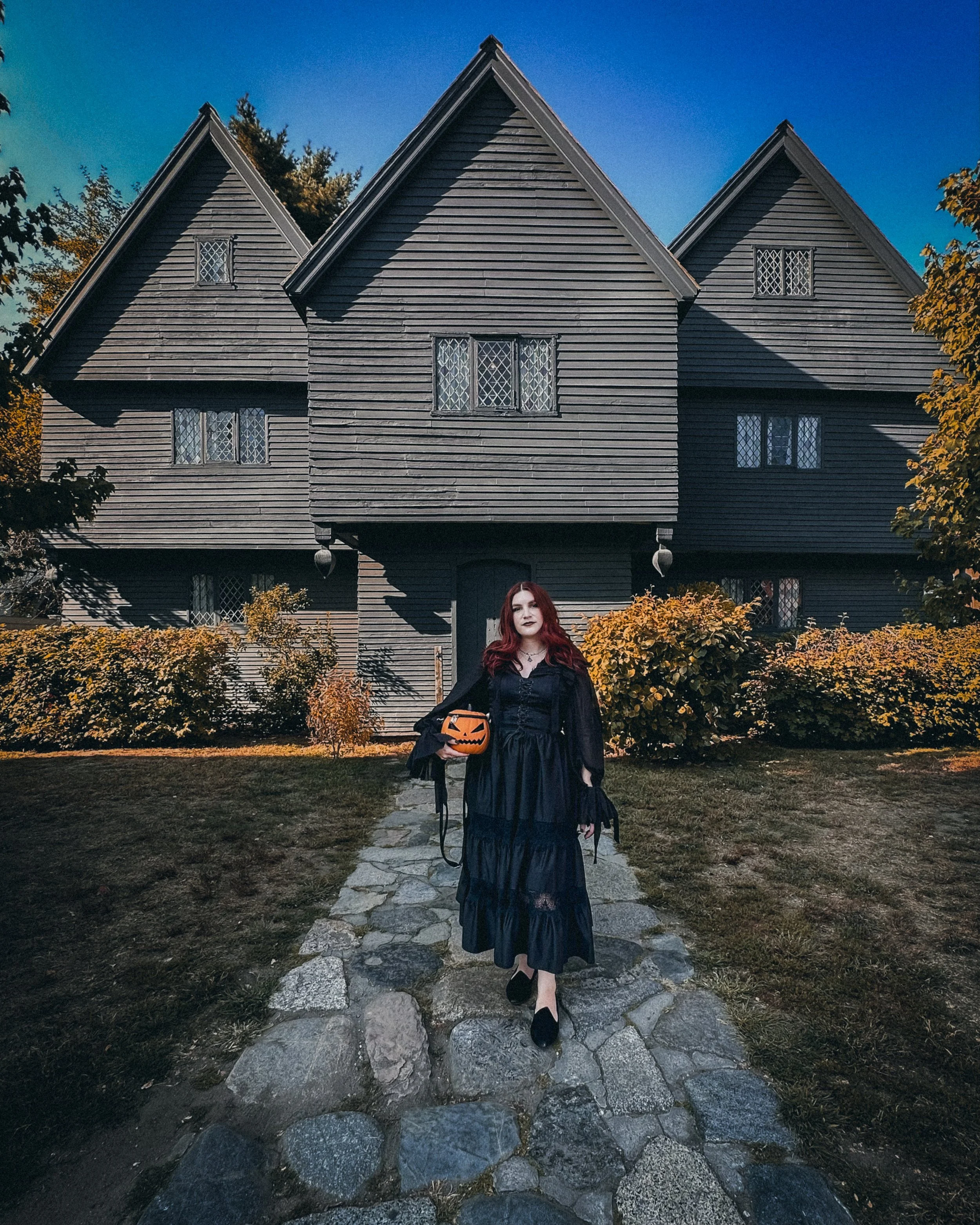 A person in a long black dress holding a pumpkin bag stands in front of a historic, dark-colored building with a stone pathway.
