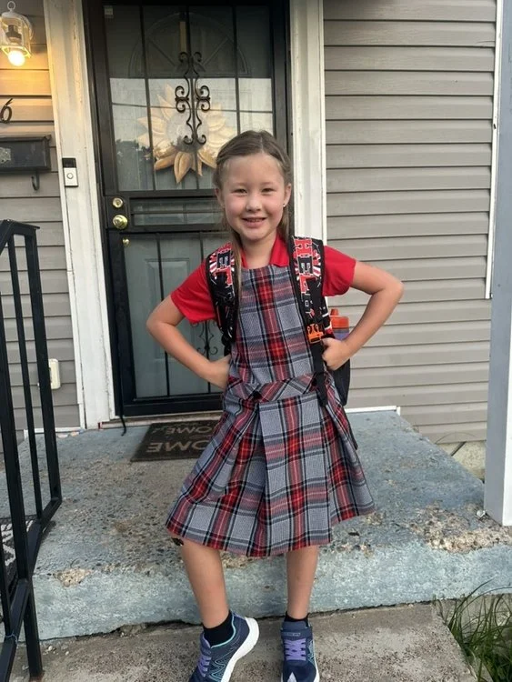 A young girl in a plaid skirt and red shirt standing on a concrete porch in front of a black door with a decorative metal window and a floral wreath. She is smiling, has her hands on her hips, and carries a backpack.