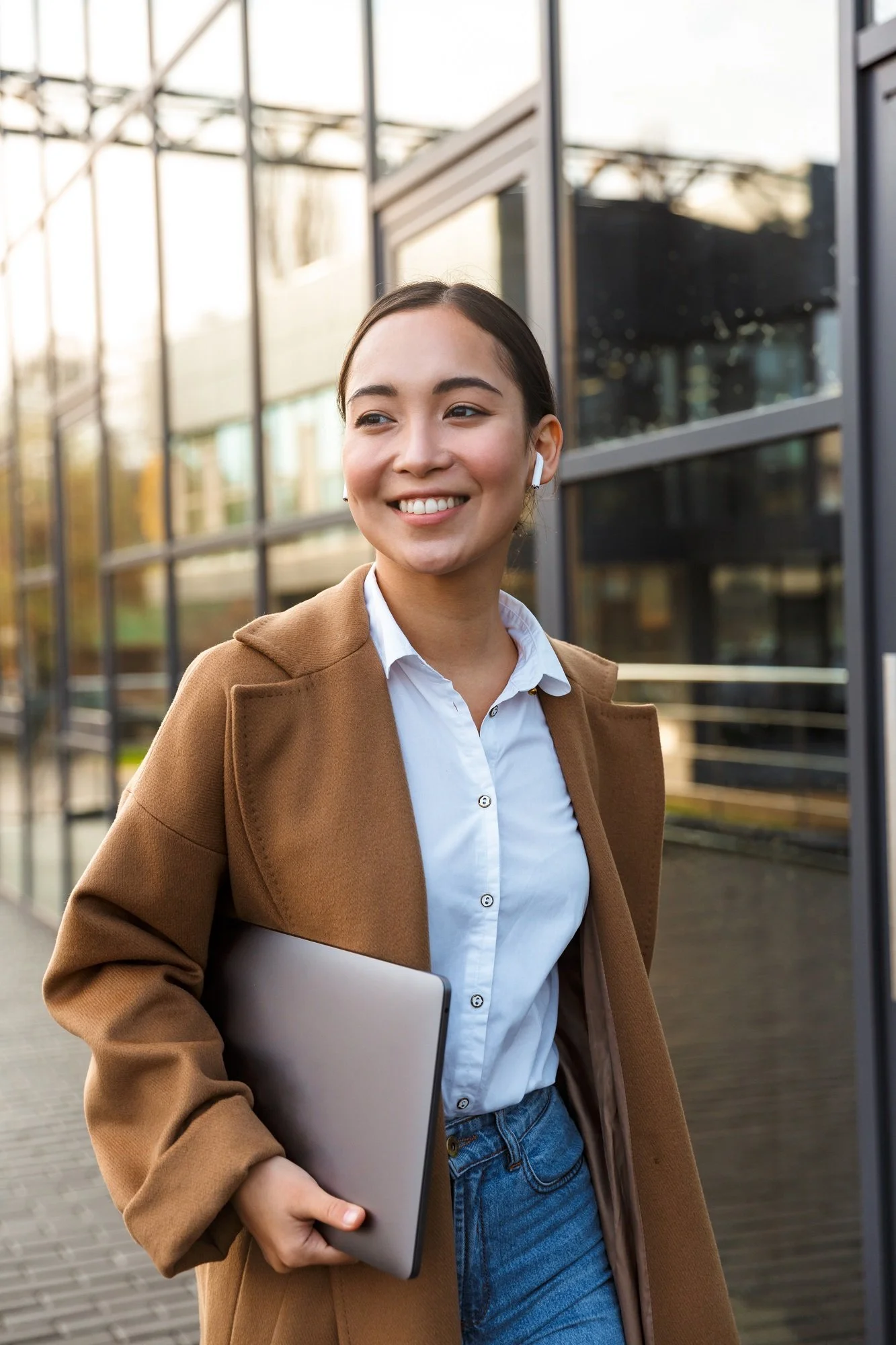 A smiling young woman with earbuds holding a laptop, standing outdoors in front of a student essay services agency.