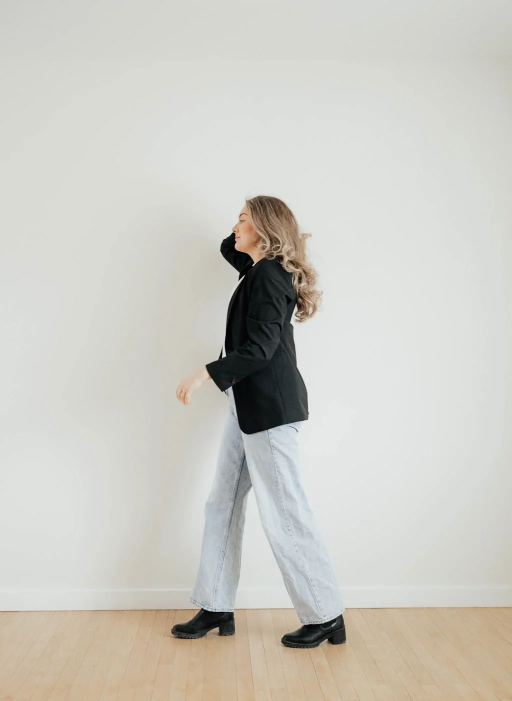 Woman with blonde hair wearing a black blazer, white top, light-colored jeans, and black ankle boots standing against a plain off-white wall.