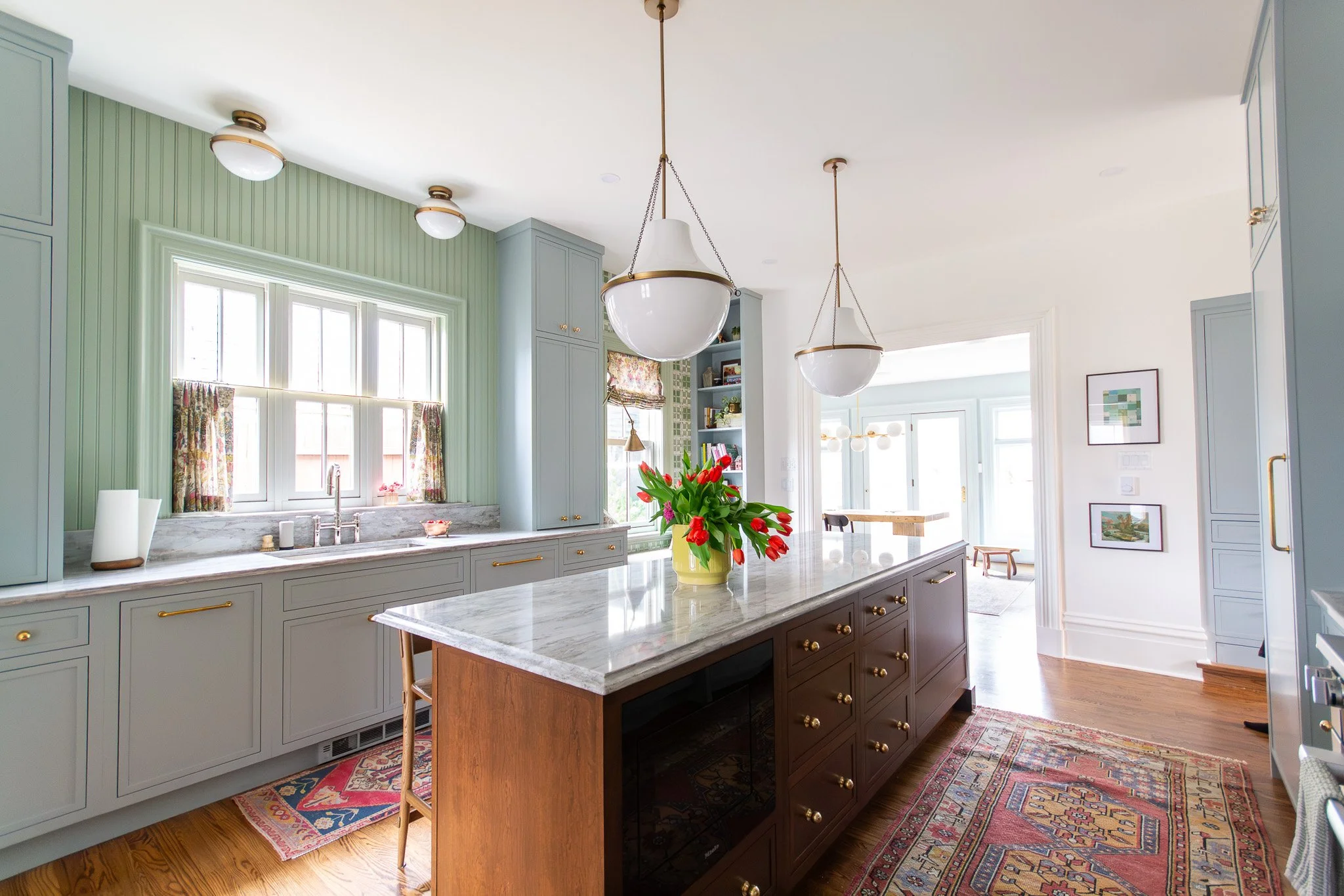 A bright kitchen with green-paneled walls and white trim. There are large windows with floral curtains, and a marble countertop with a sink. A bouquet of red flowers in a yellow vase sits on a white marble island with a dark wood base and brass hardware. Pendant lights hang from the ceiling, and there is a colorful rug on the wooden floor.