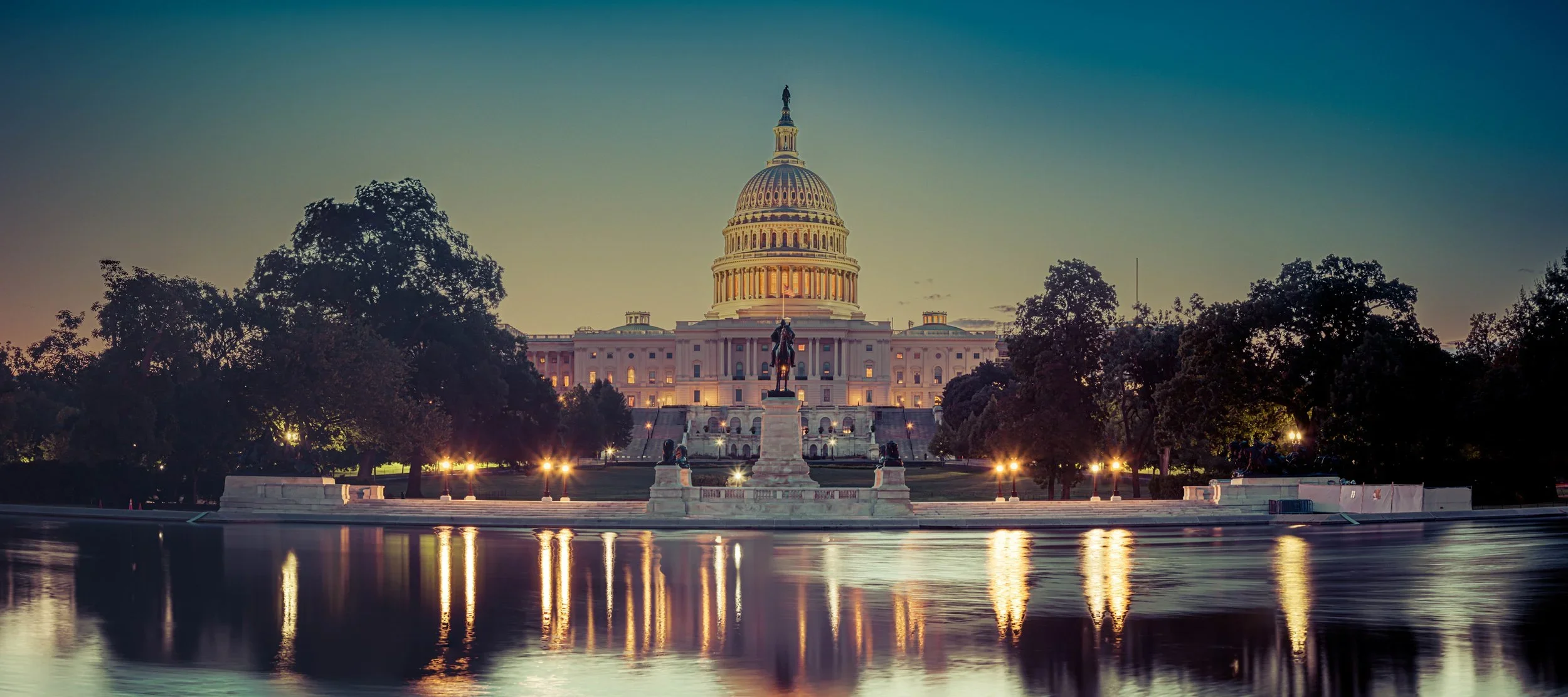 The U.S. Capitol building illuminated at sunset, reflecting in the water of the reflecting pool in Washington, D.C., with trees surrounding it.