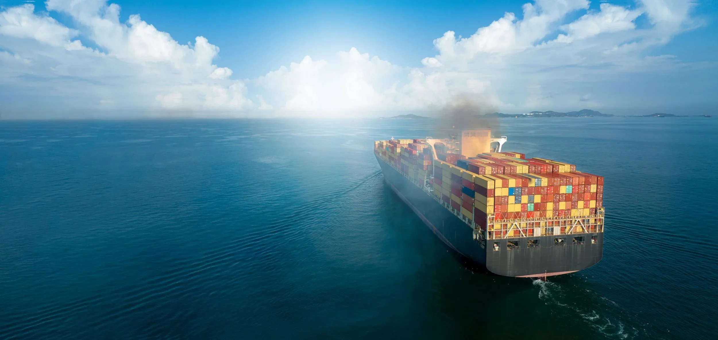 A large cargo ship carrying stacked shipping containers sailing through blue ocean waters with distant islands on the horizon and a partly cloudy sky.