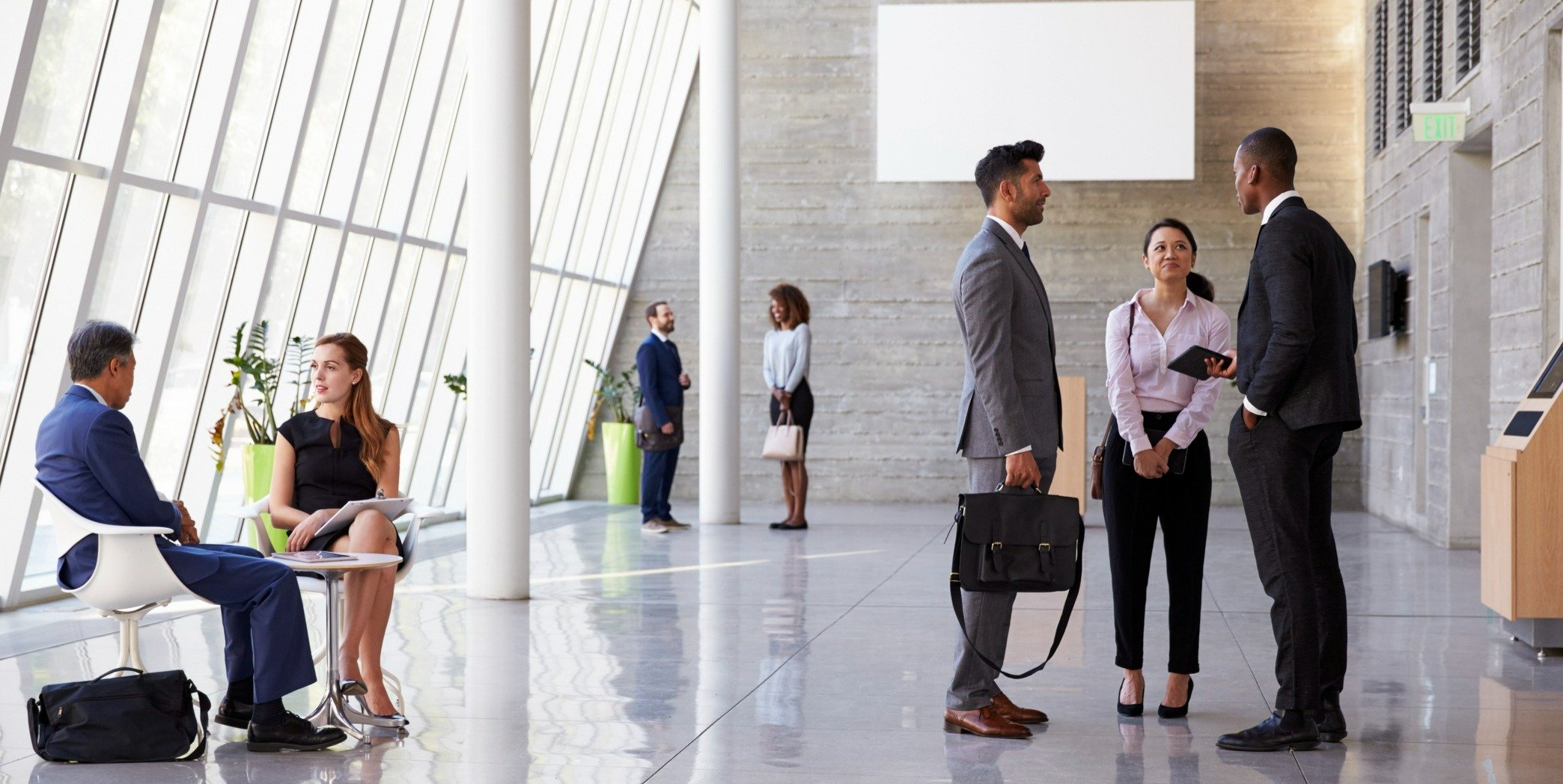 Modern office lobby with professionals engaged in conversations, dressed in business attire, with large windows allowing natural light to fill the space.