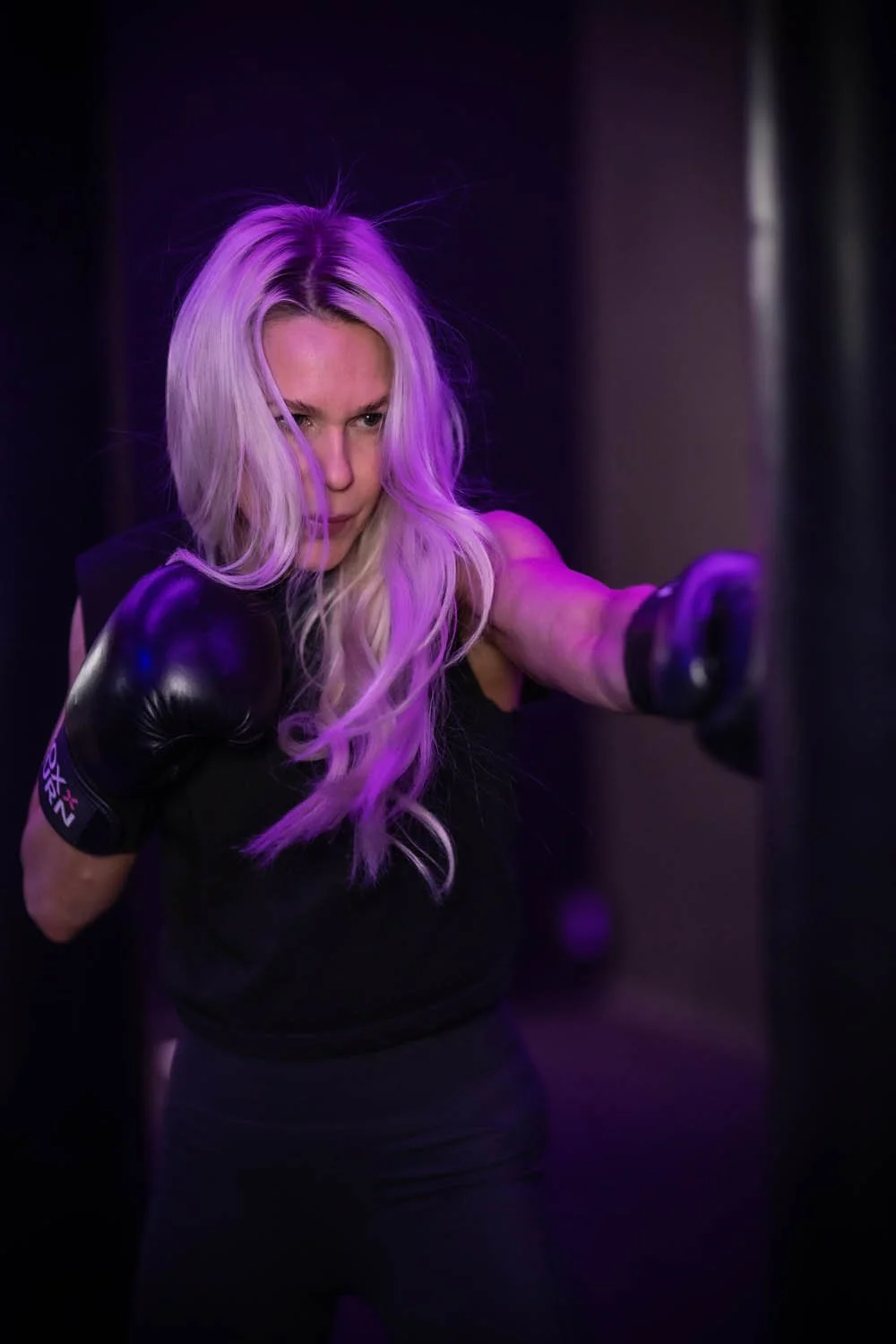 Nainen treenaa nyrkkeilyä pimeässä tilassa, valaistuksessa violetti valo.