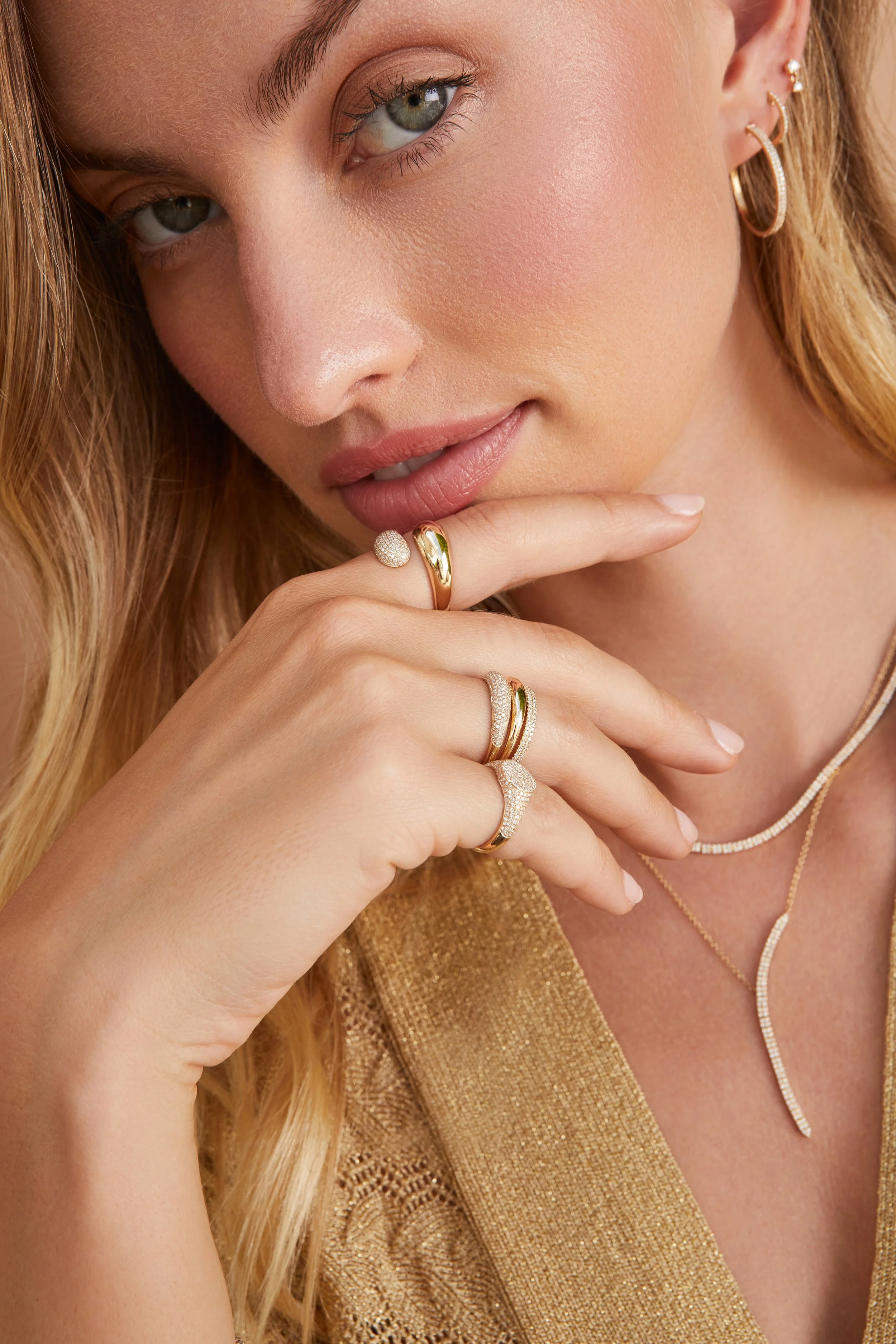 Close-up of a woman with blonde hair wearing gold jewelry, including multiple rings, earrings, and layered necklaces, with a gold-colored top.