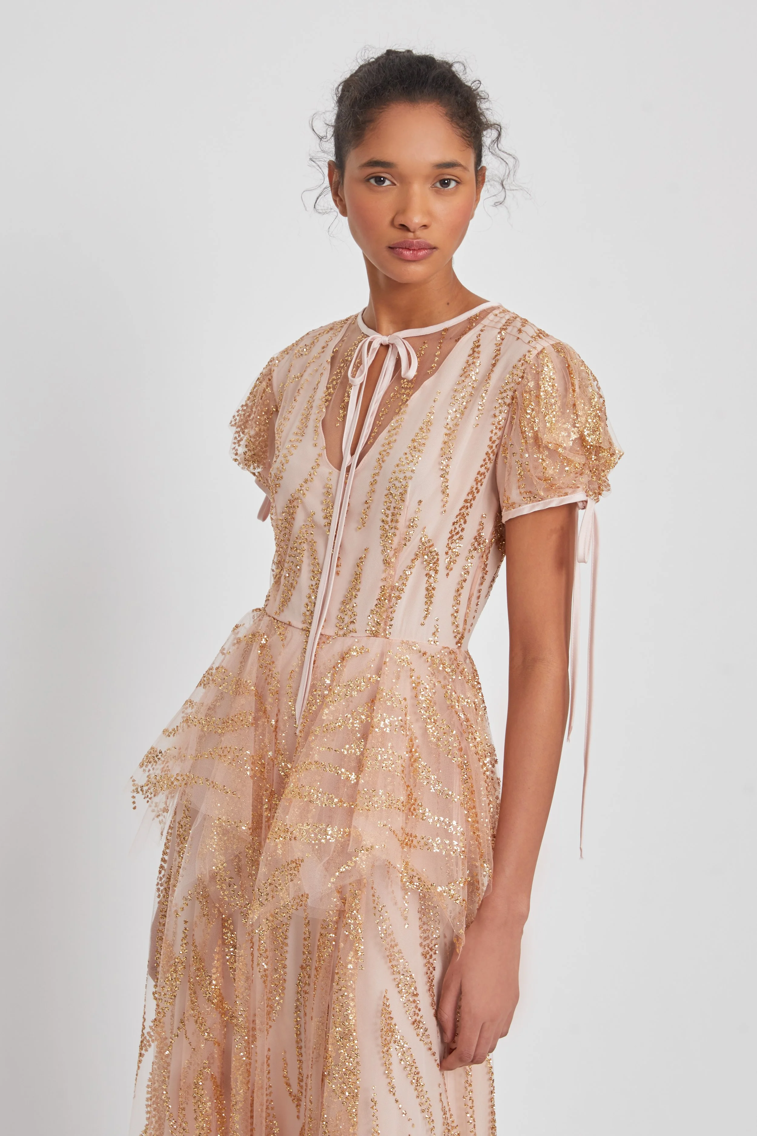 A young woman wearing a pink and gold glittery dress with puffed sleeves and a tie at the neckline, standing against a plain background.