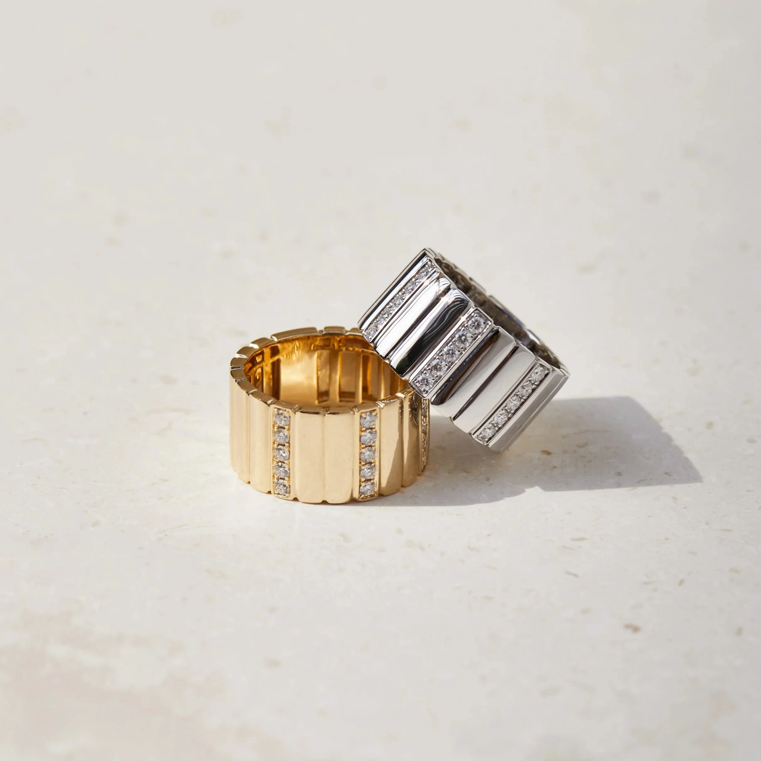 Two stacked rings, one gold with diamonds and one silver with diamonds, on a light-colored surface.