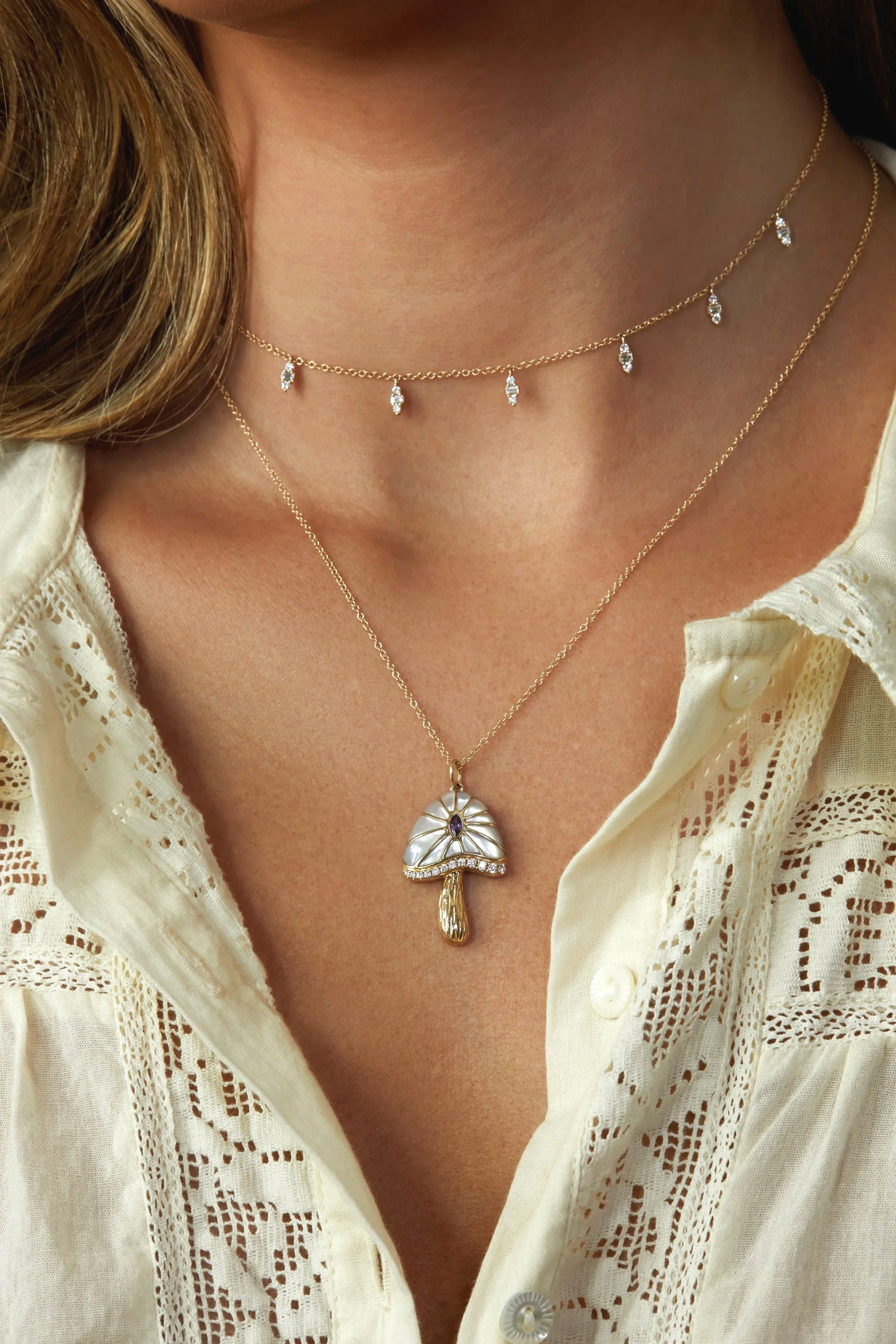 Close-up of a woman wearing layered gold necklaces, one with small diamond-like charms and the other with a mushroom-shaped pendant with a purple stone, over a cream-colored lace blouse.