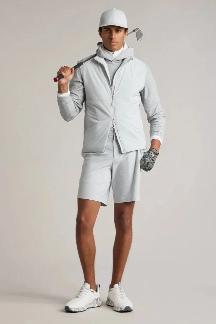 Young man dressed in golf attire, holding a golf club over his shoulder, wearing a white cap, vest, shorts, and sneakers.