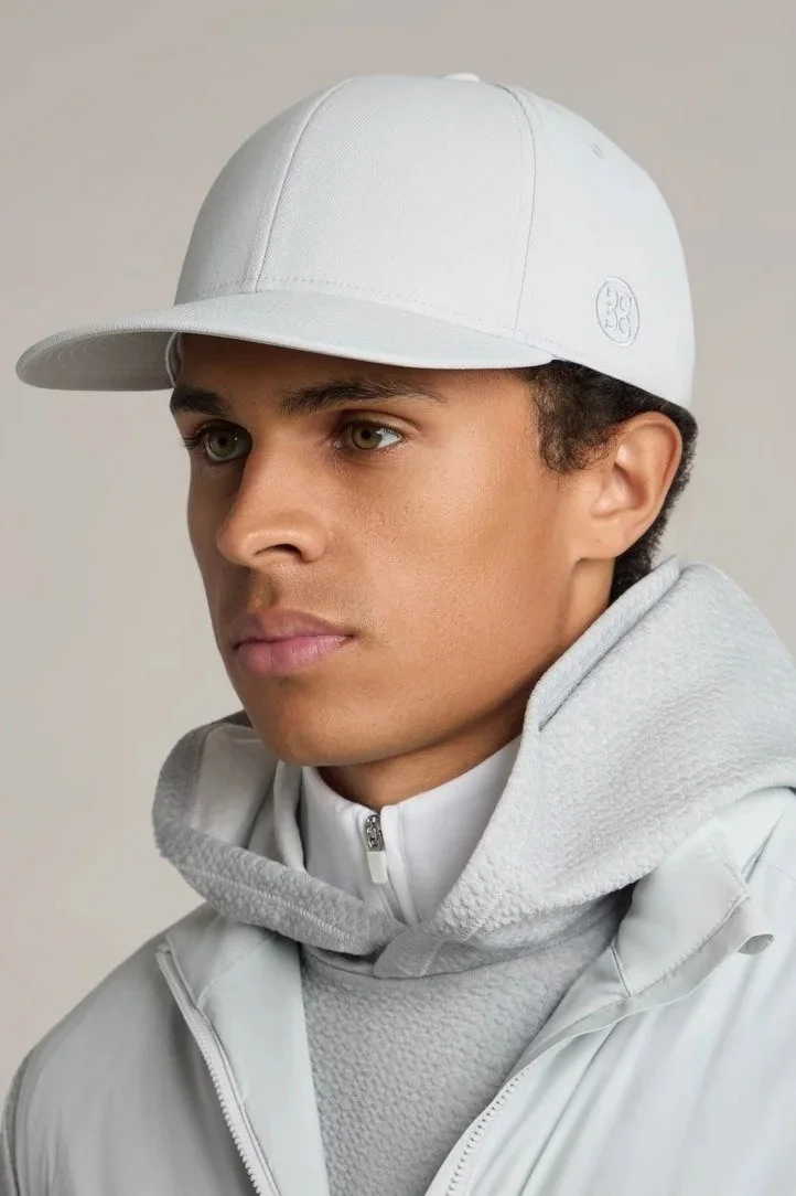 Young man wearing a white baseball cap, white jacket, and gray hoodie, looking to the side against a plain background.