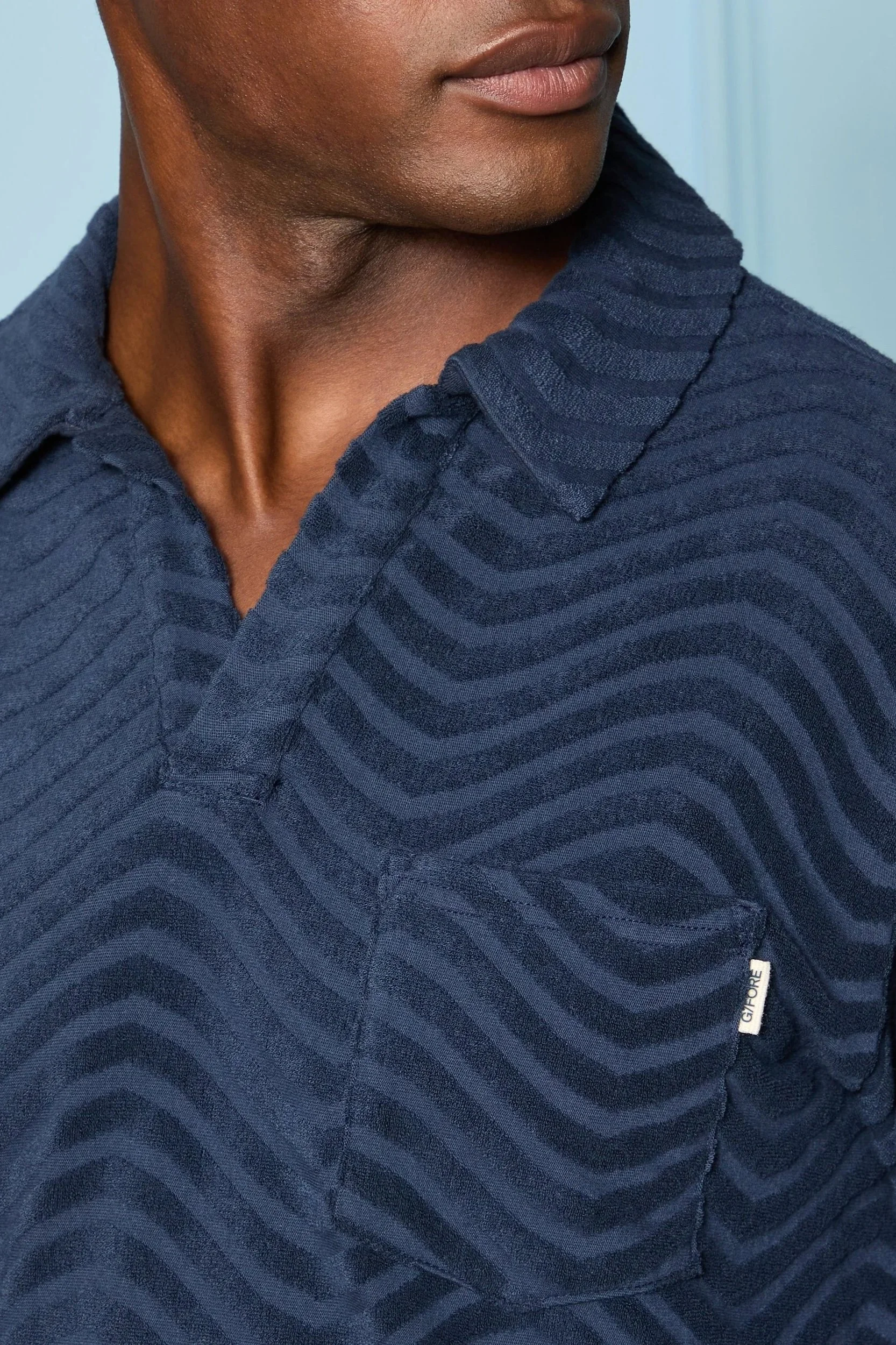 Close-up of a person wearing a navy blue shirt with a wave-like pattern, showing part of their face and shoulder.