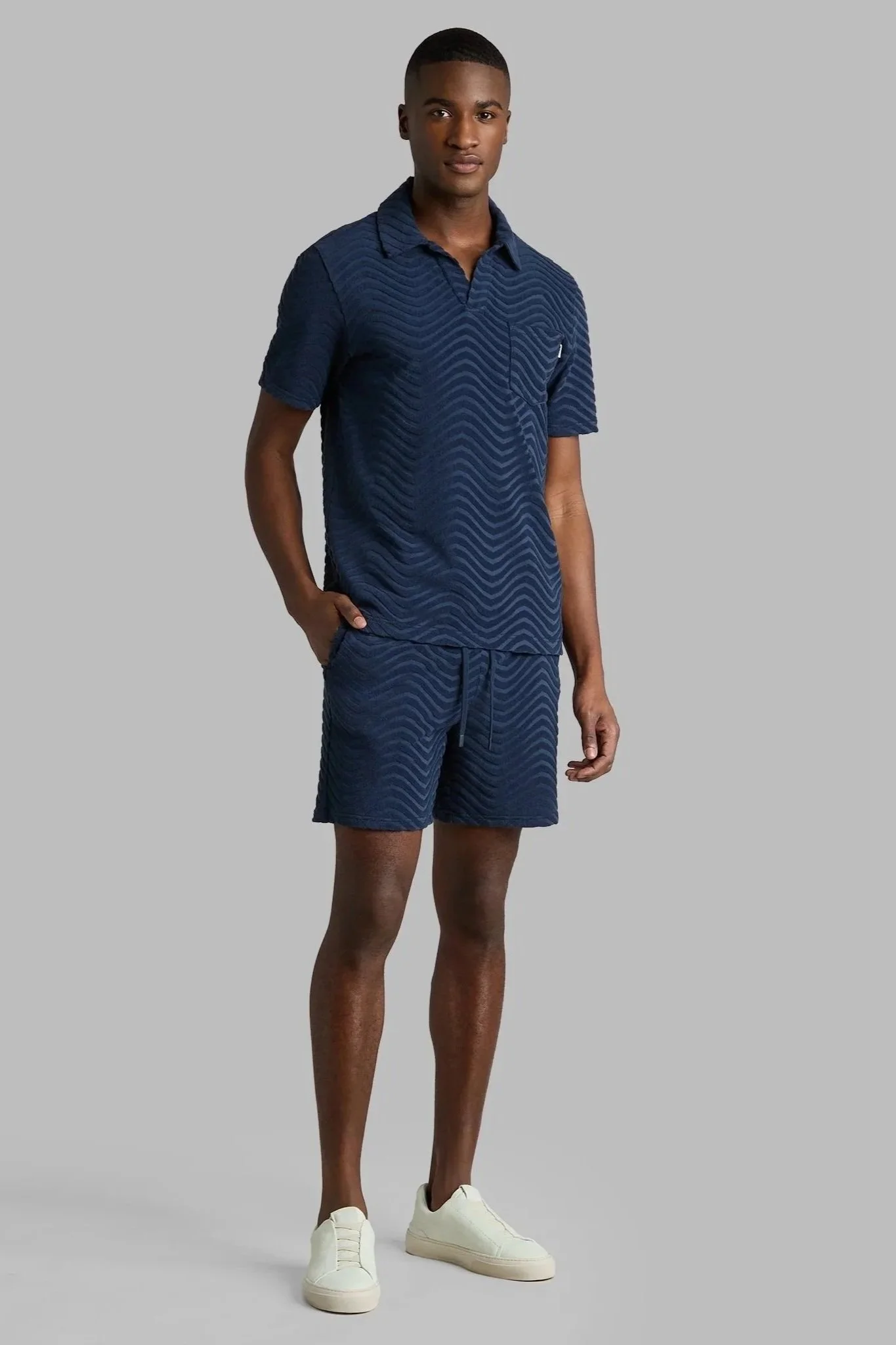 Young man in navy blue athletic shirt and shorts, white sneakers, standing against gray background.