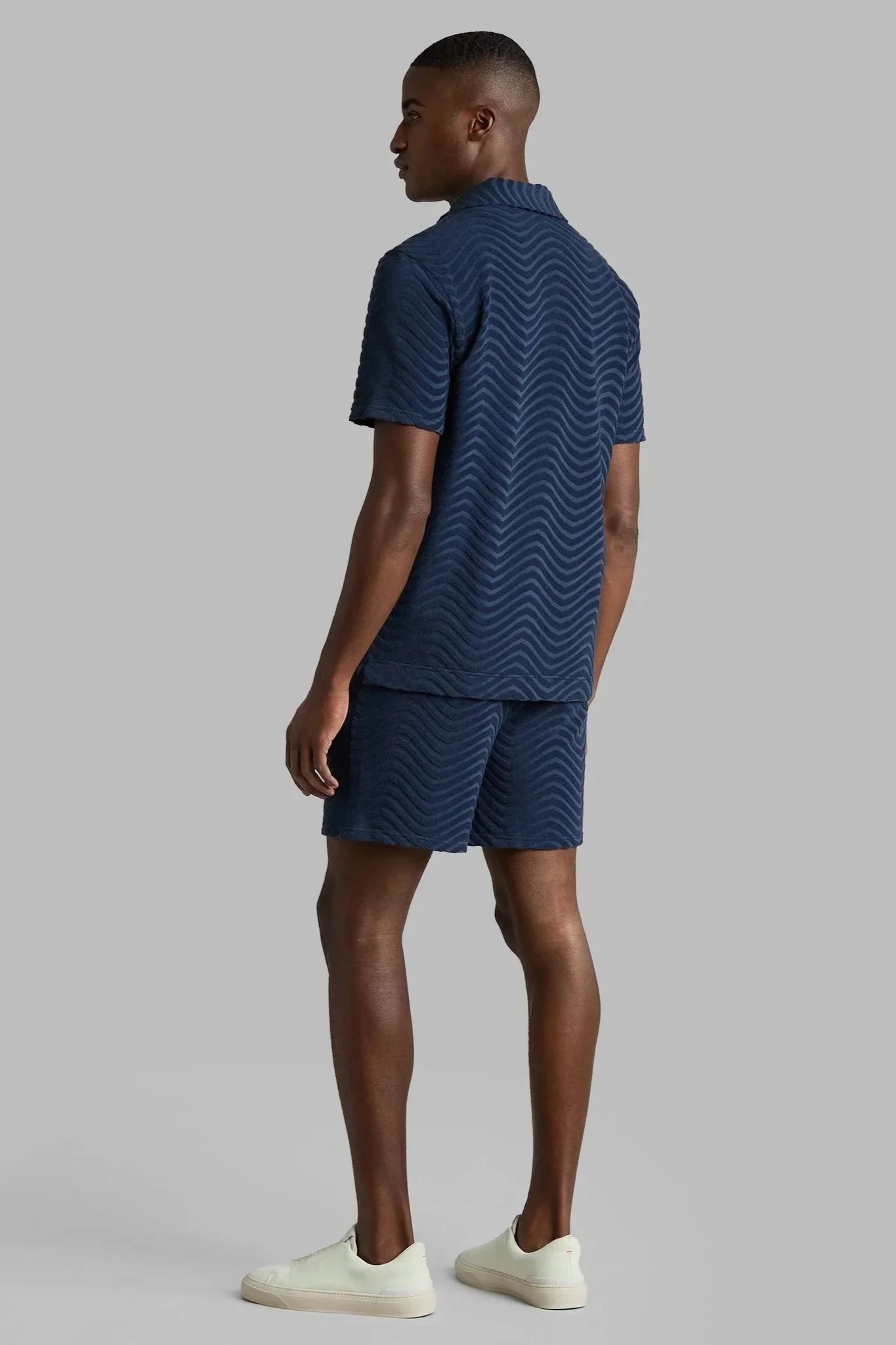 A man standing with his back to the camera, wearing a navy blue textured short-sleeve shirt, matching shorts, and white sneakers, against a plain gray background.