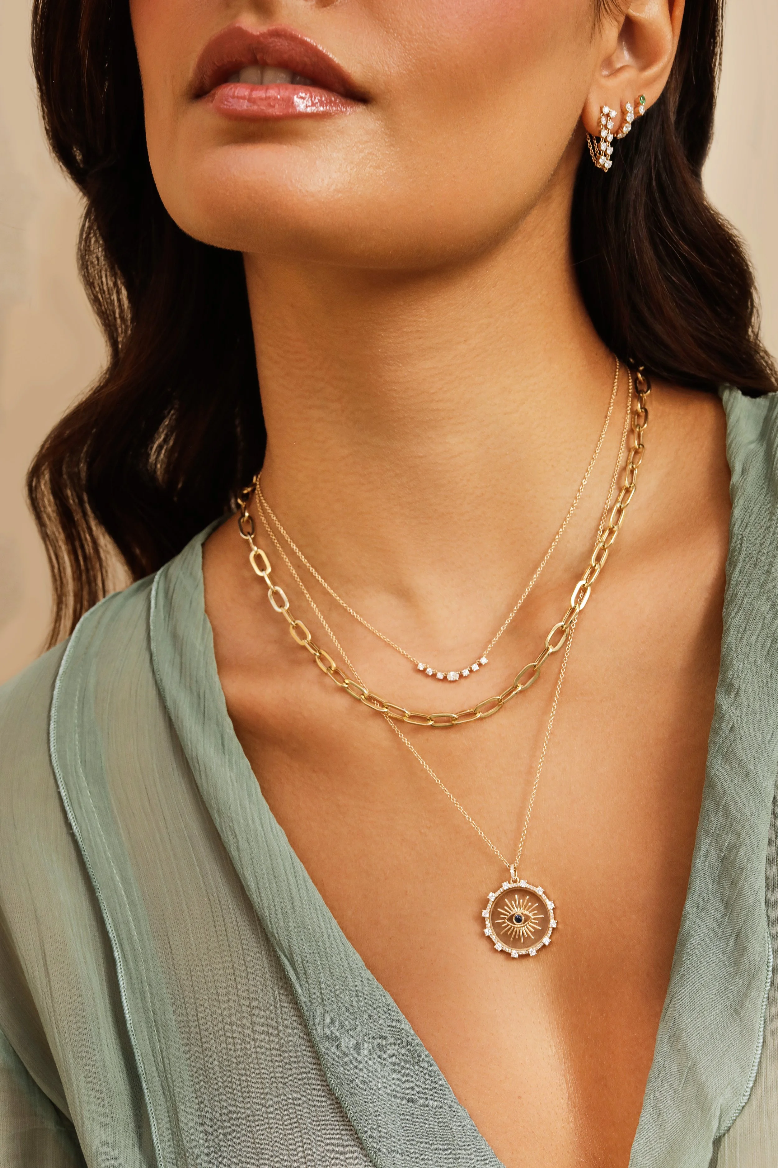 Close-up of a woman's neck and lower face showing layered gold necklaces, including a chain with small diamonds, a chain with a circular pendant featuring an eye and sunburst design, and a chain with large links. She is wearing a light green blouse w
