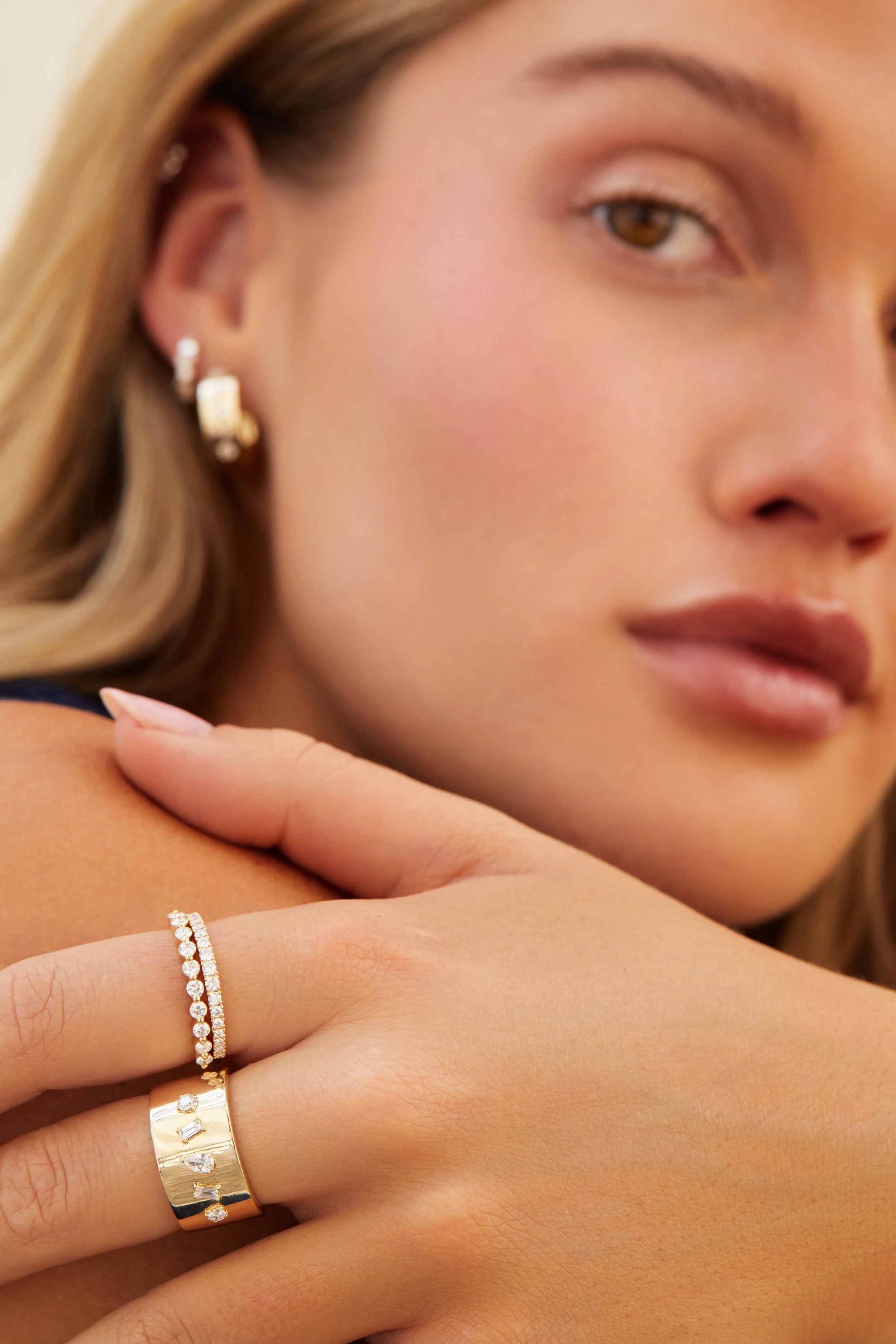Close-up of a woman with gold earrings and rings, touching her shoulder.