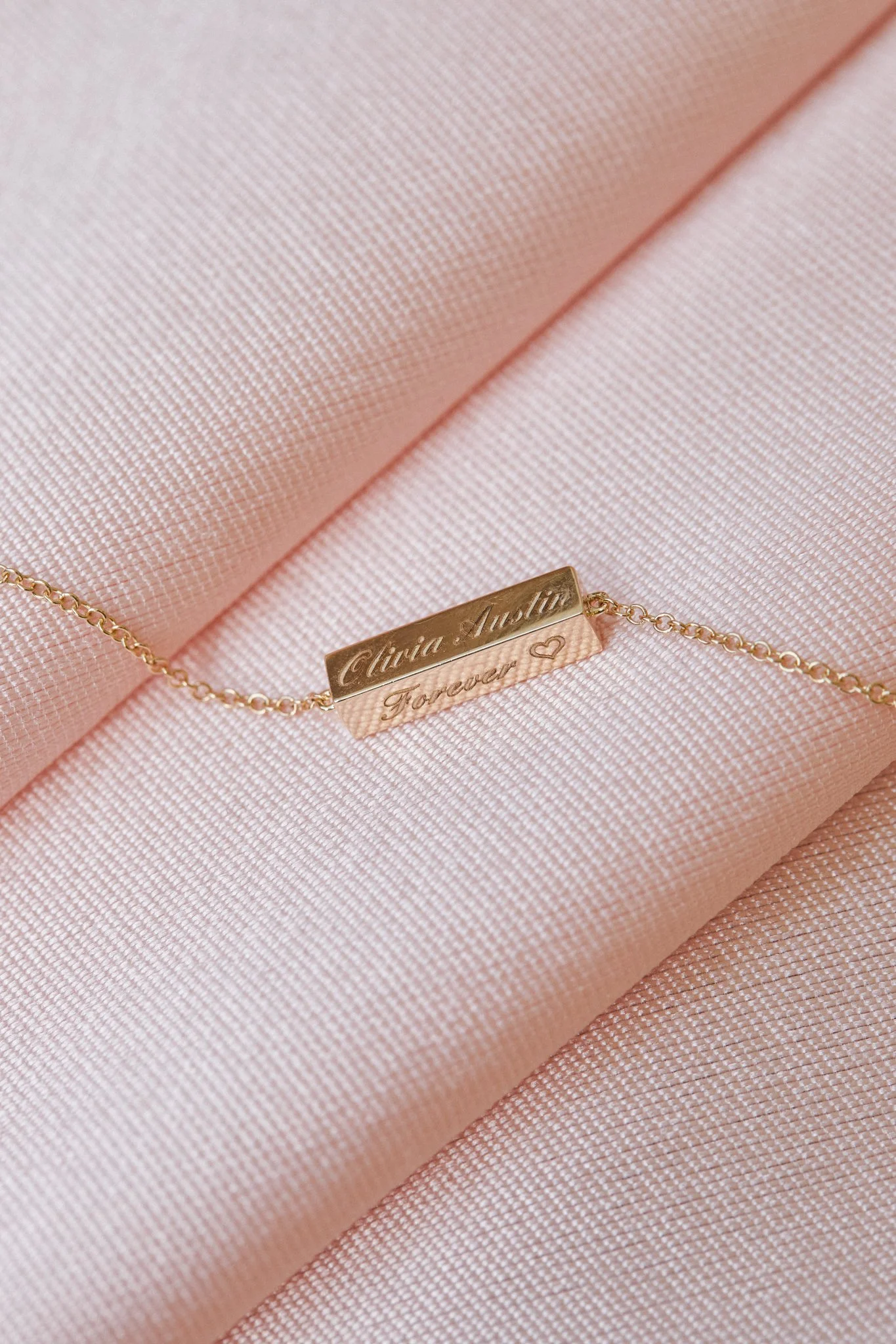 Gold necklace with a rectangular pendant engraved with the names 'Alicia Austin' and the words 'Forever' with a small heart, resting on pink fabric.