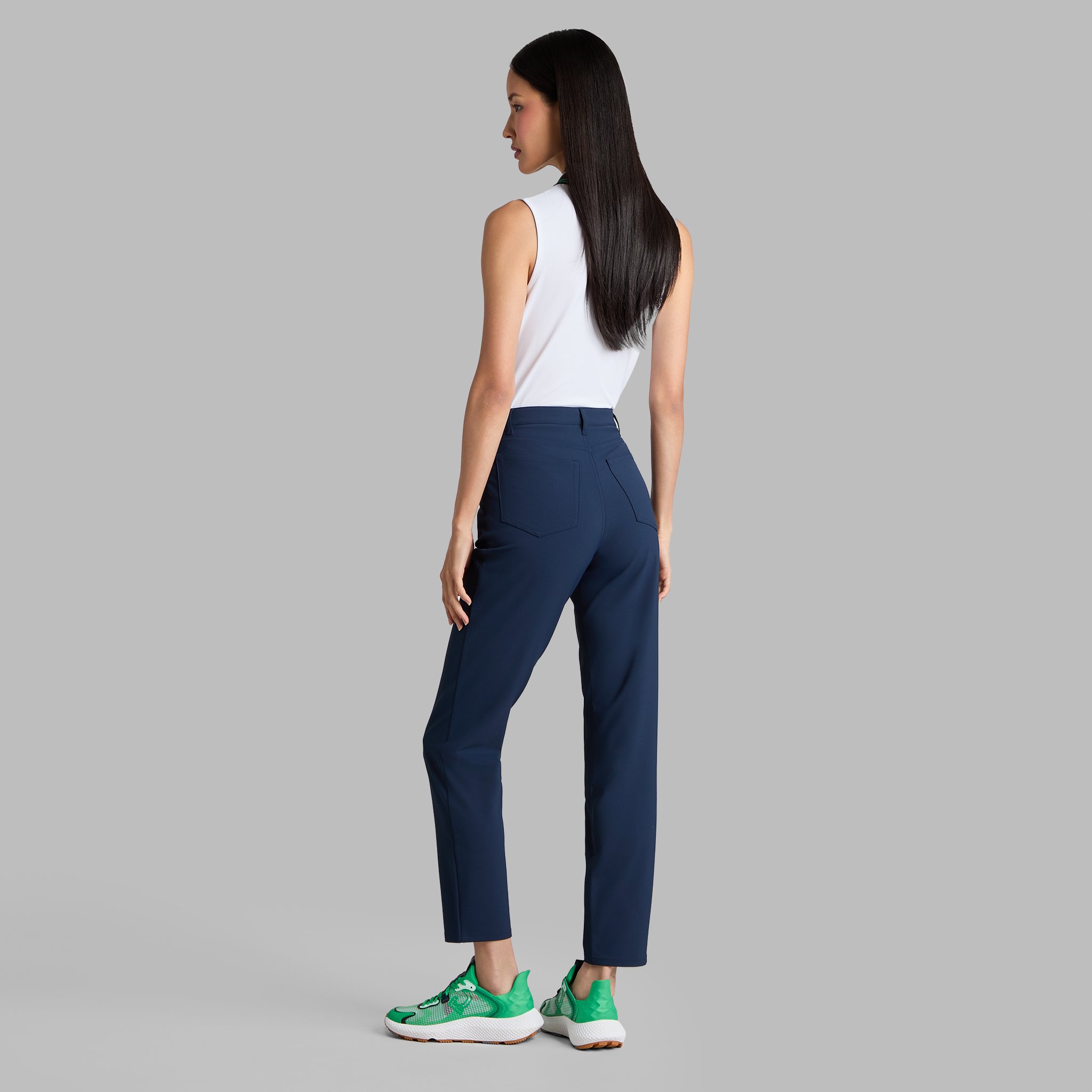 A woman with long dark hair in a white sleeveless top, navy blue pants, and green sneakers standing against a plain gray background, facing away.