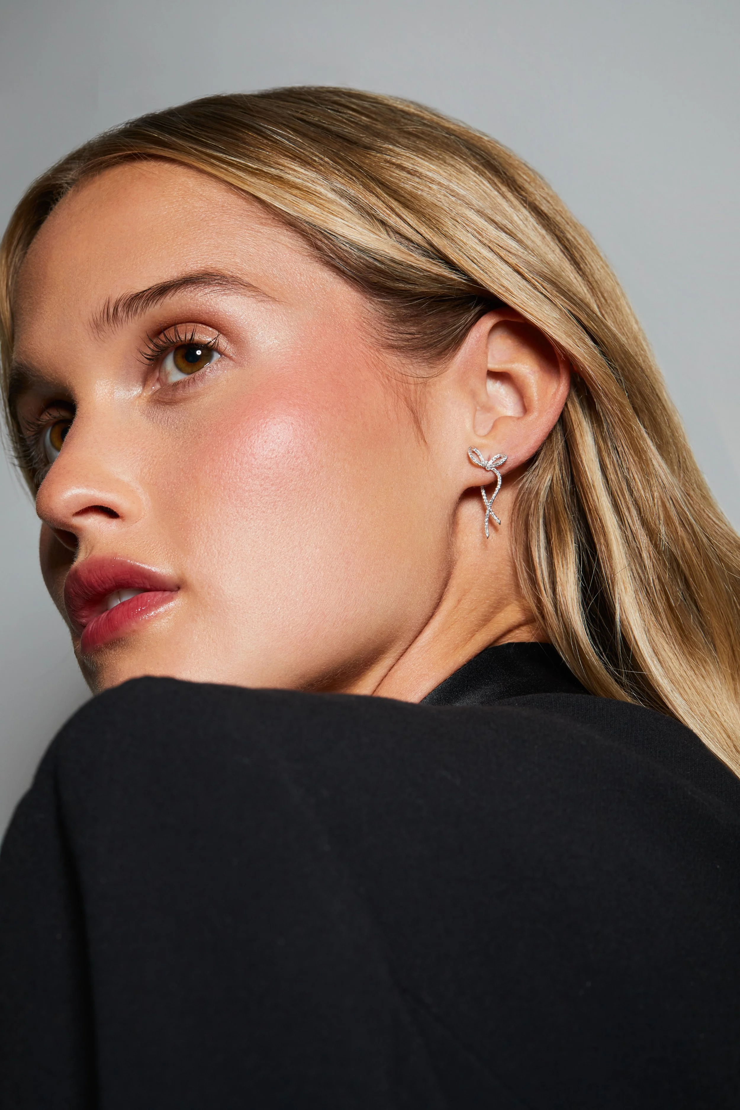 Close-up of a woman with blonde hair and makeup, wearing a jewelry earring with a leaf-like design, against a neutral background.