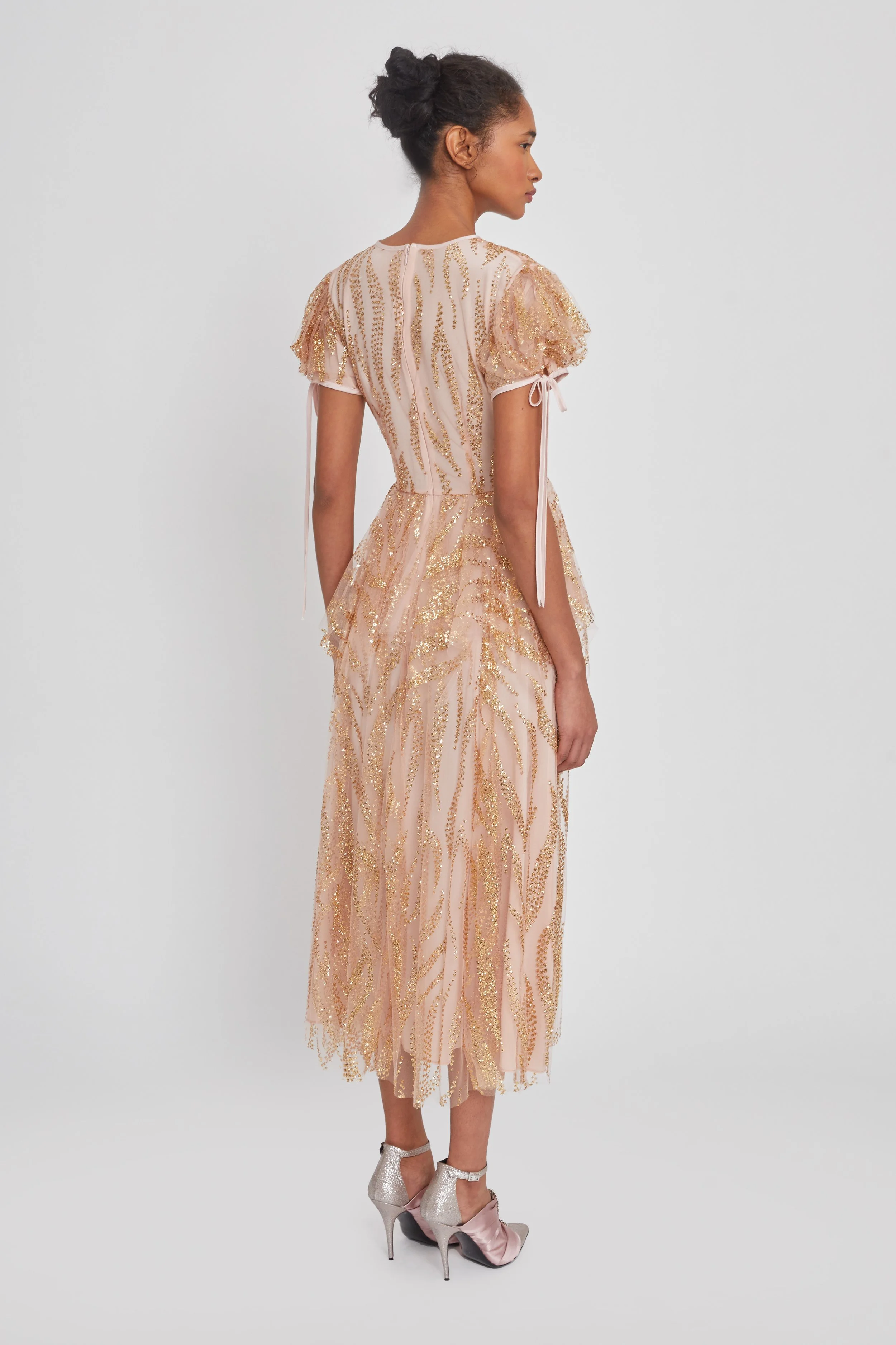 A woman in a pink and gold beaded dress with short puffed sleeves and matching high-heeled shoes, standing sideways against a plain white background.