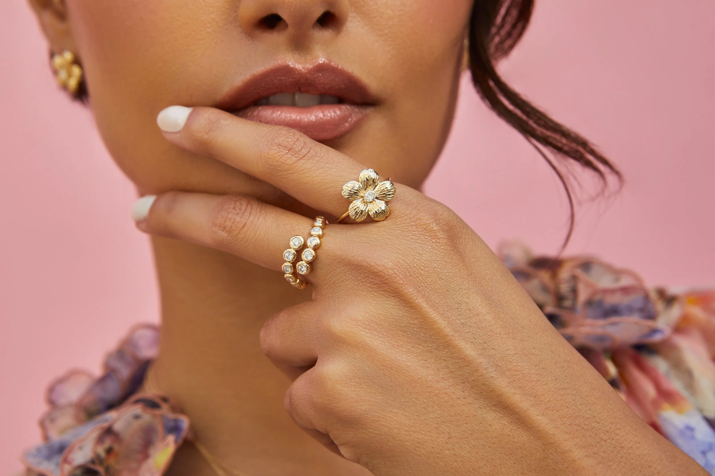 Close-up of a woman's hand wearing a flower-shaped ring and a diamond-studded ring, with her lips slightly parted, against a pink background.